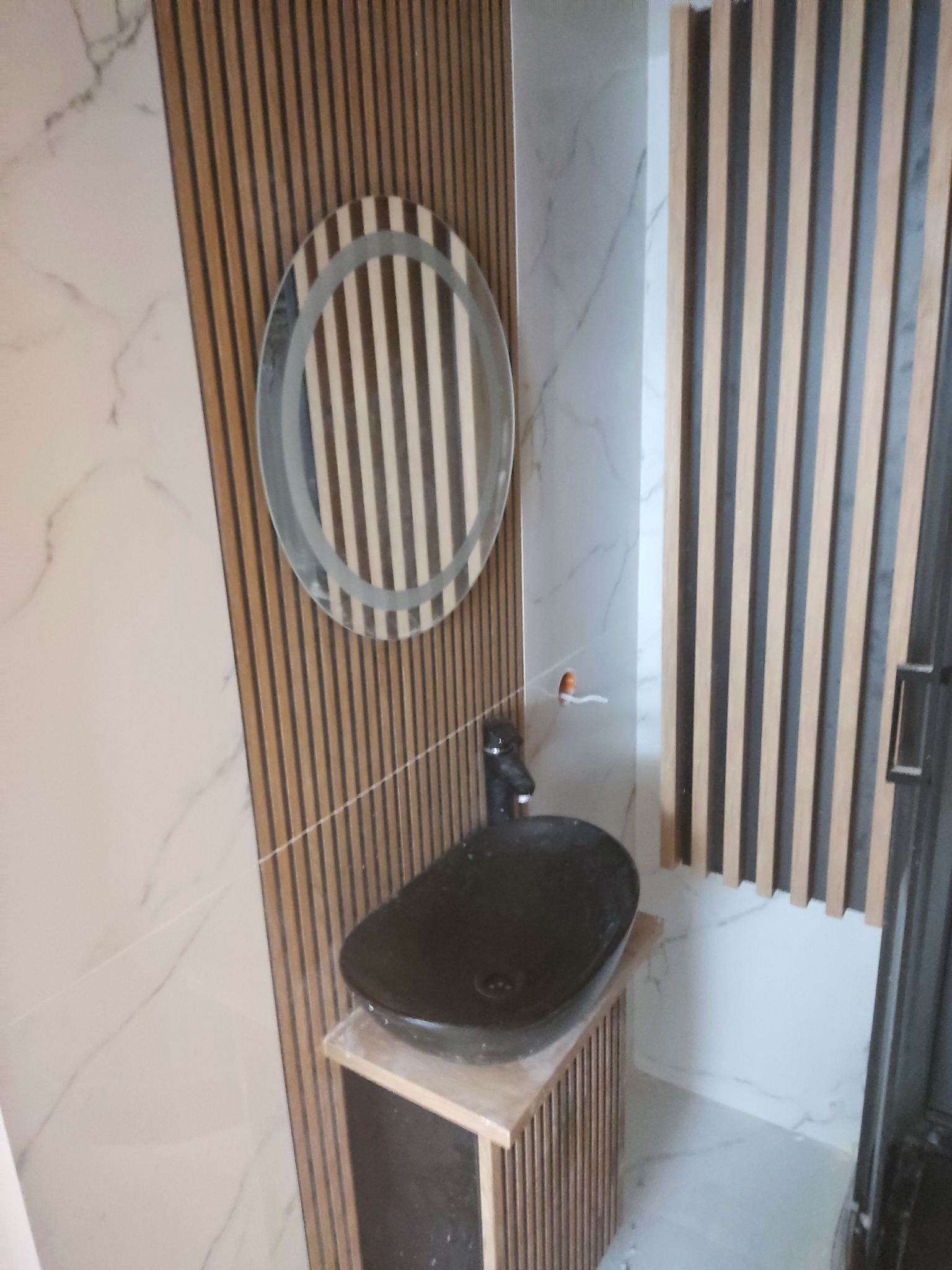 Nowoczesna łazienka z okrągłym lustrem z podświetleniem, czarną umywalką nablatową i dekoracyjnymi lamelami na ścianie, wykończona płytkami imitującymi marmur.