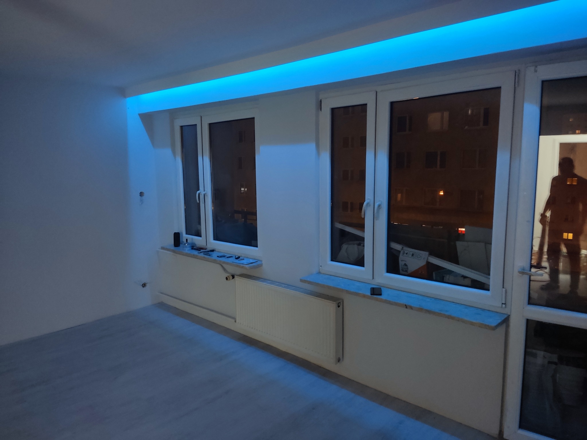 Białe wnętrze z oknami, grzejnikami i niebieskim oświetleniem LED podsufitowym. Widok na nocne miasto przez okna. Świeżo wykończone, puste pomieszczenie.