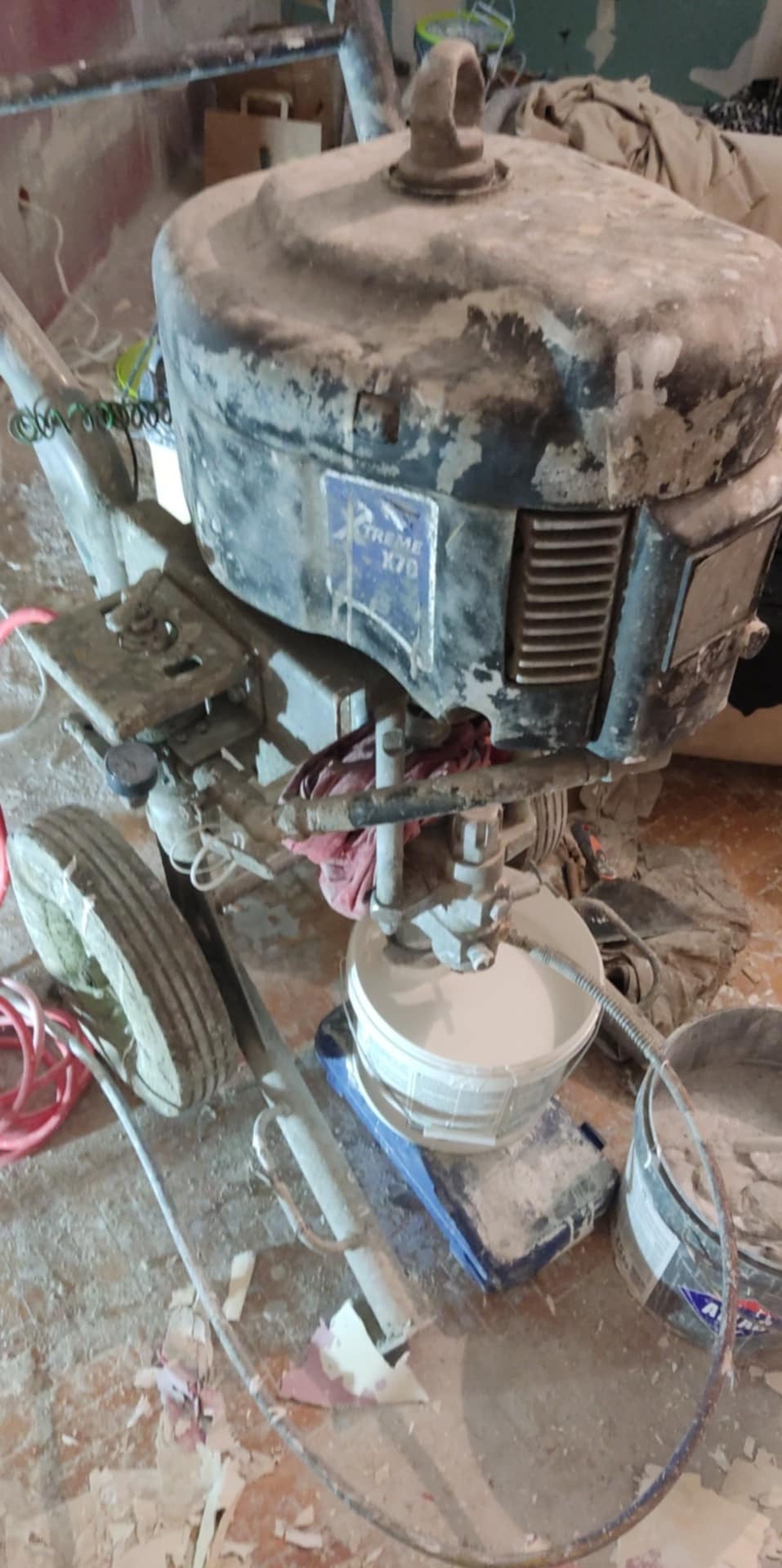 Brudny agregat malarski Xtreme X70 z wiadrem farby w pomieszczeniu w trakcie prac wykończeniowych. Widoczne ślady farby i pyłu.