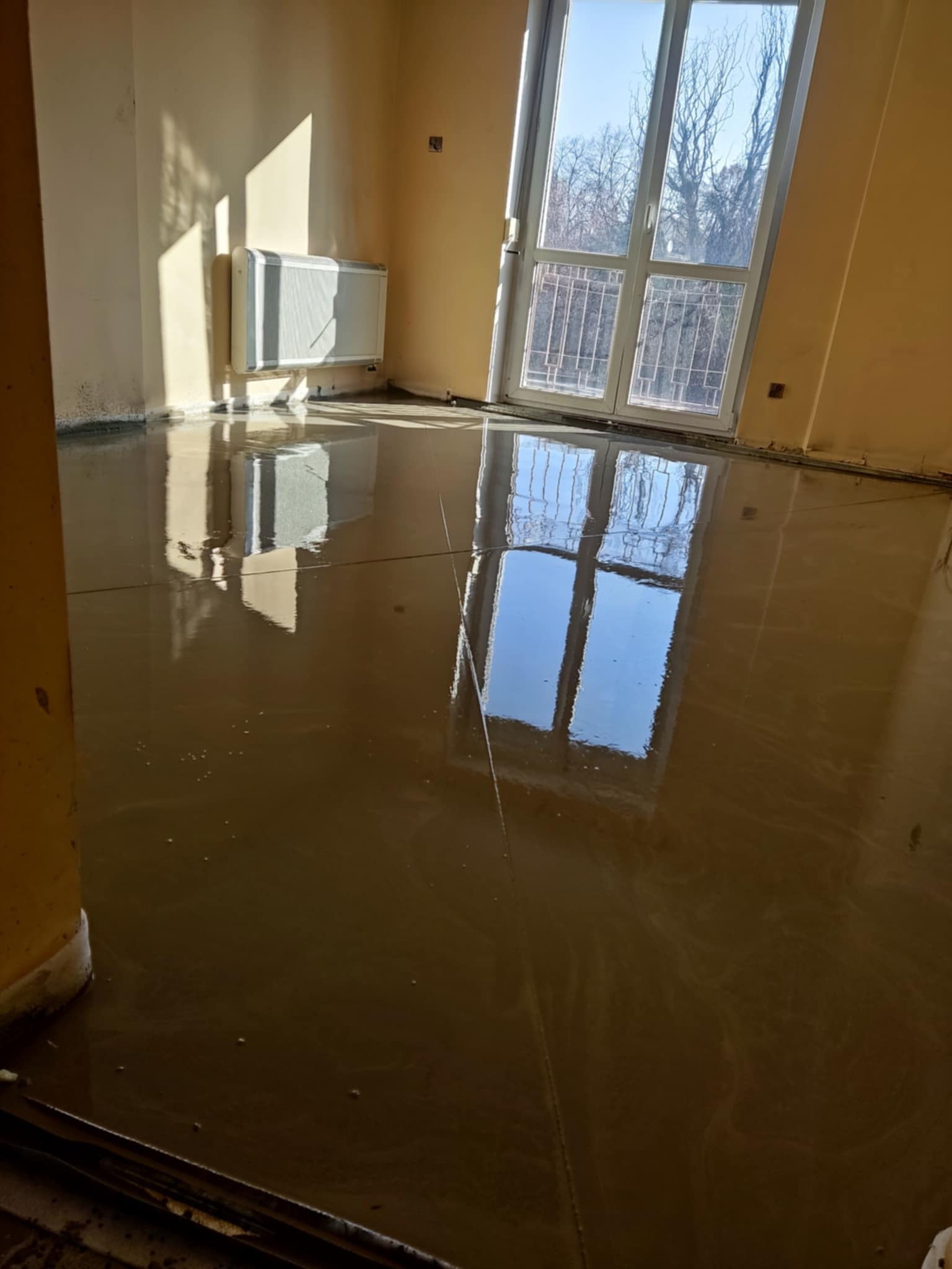 Wylana, mokra wylewka samopoziomująca w pokoju z grzejnikiem i oknem. Widoczne odbicie okna w mokrej powierzchni. Ściany w kolorze żółtym.