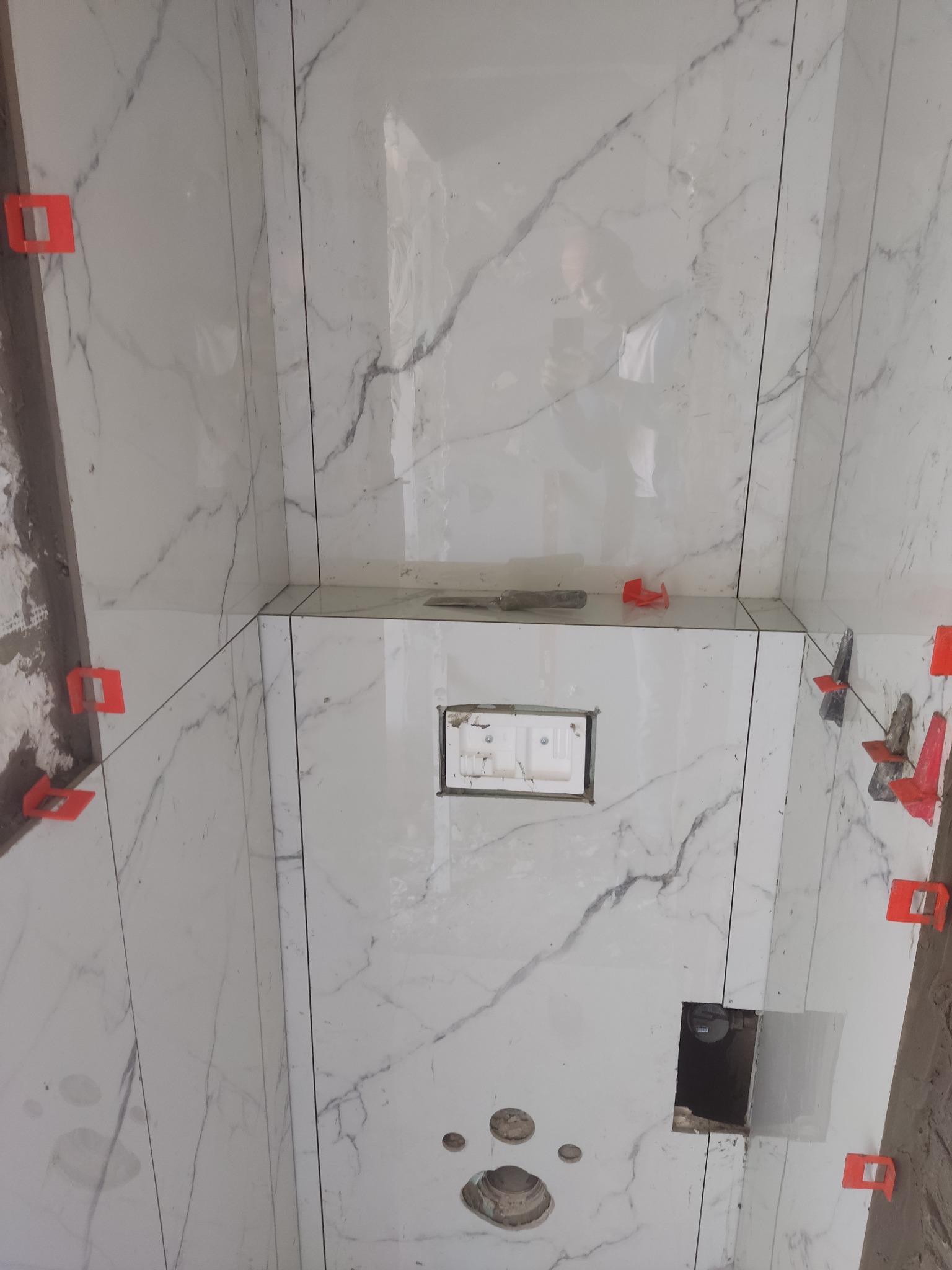 Białe płytki imitujące marmur z czerwonymi klipsami poziomującymi, zamontowane w łazience. Widoczne otwory na instalacje i odbicie osoby robiącej zdjęcie.