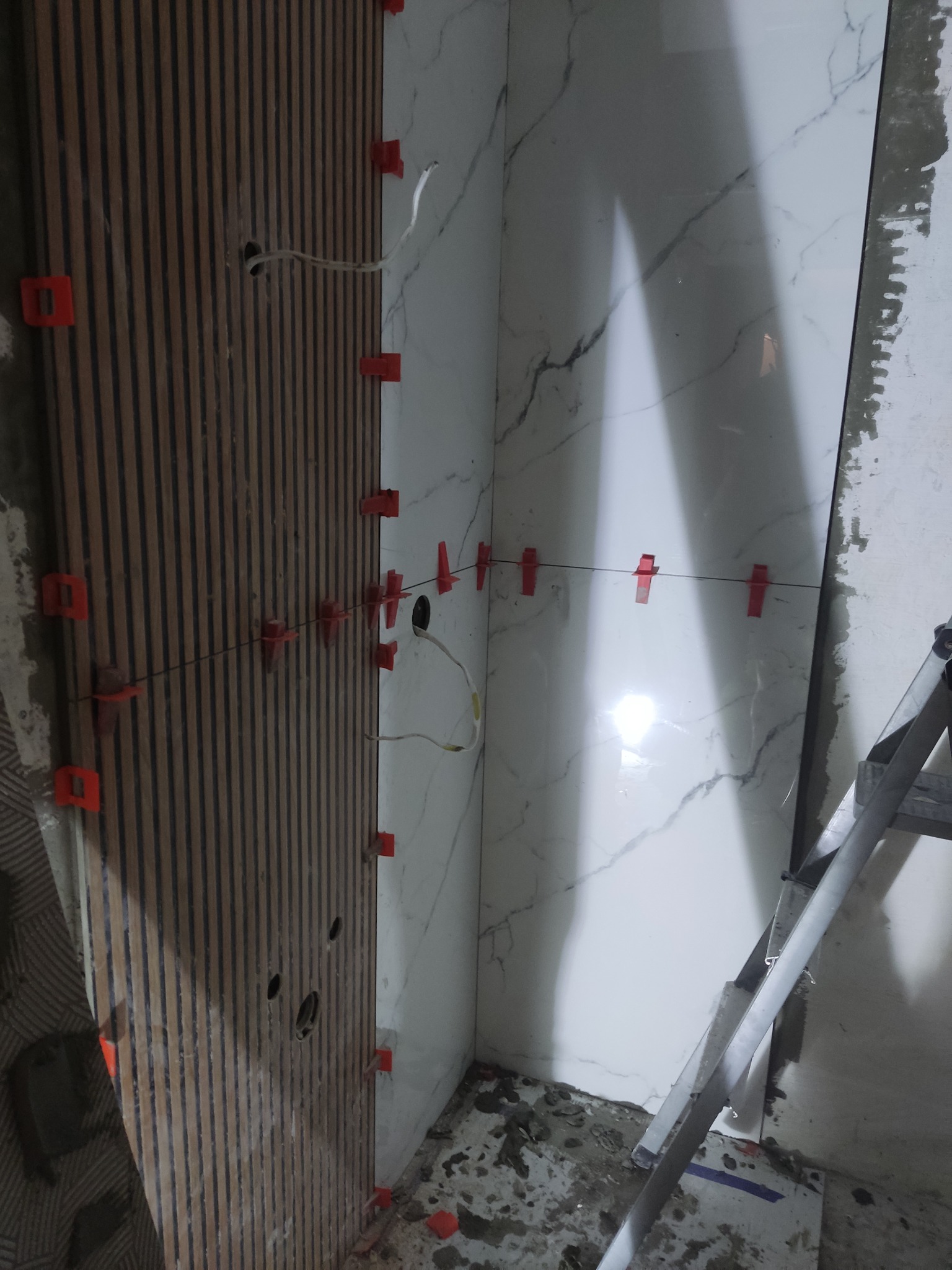 Układanie płytek imitujących marmur i drewnianych paneli ściennych z użyciem klipsów poziomujących. Widoczna drabina i przewody elektryczne.