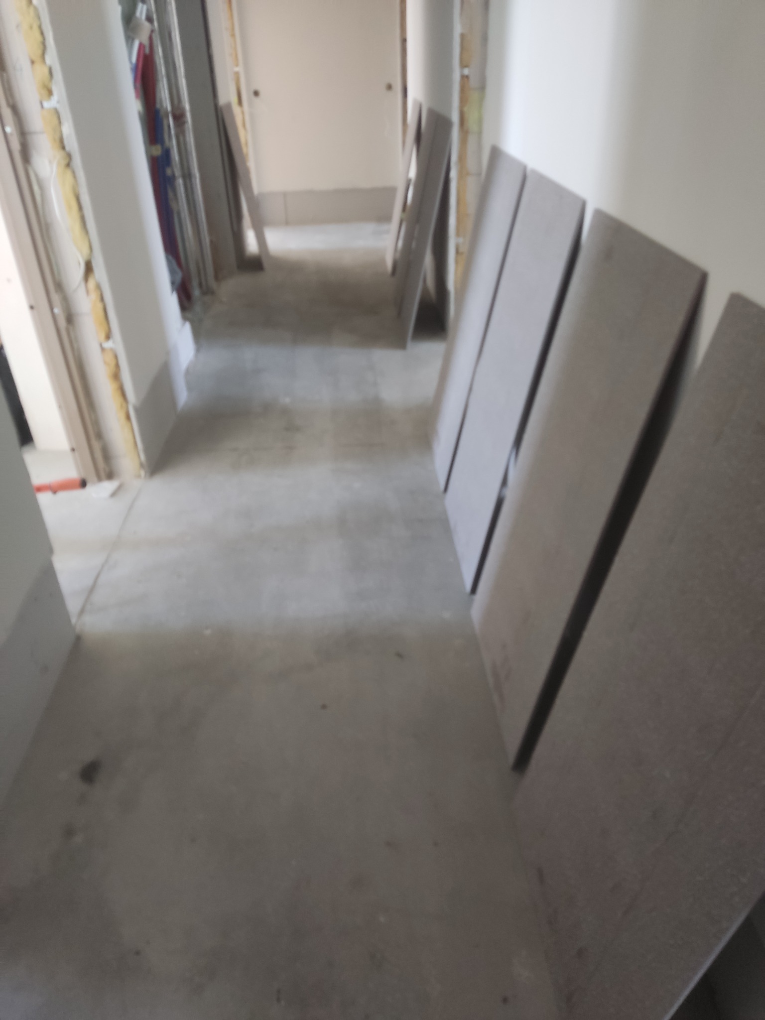 Wnętrze w trakcie remontu: widoczne płyty karton-gips o szarej fakturze, ustawione wzdłuż ściany, instalacje w ścianach, surowy beton na podłodze. Perspektywa długa, wąska.