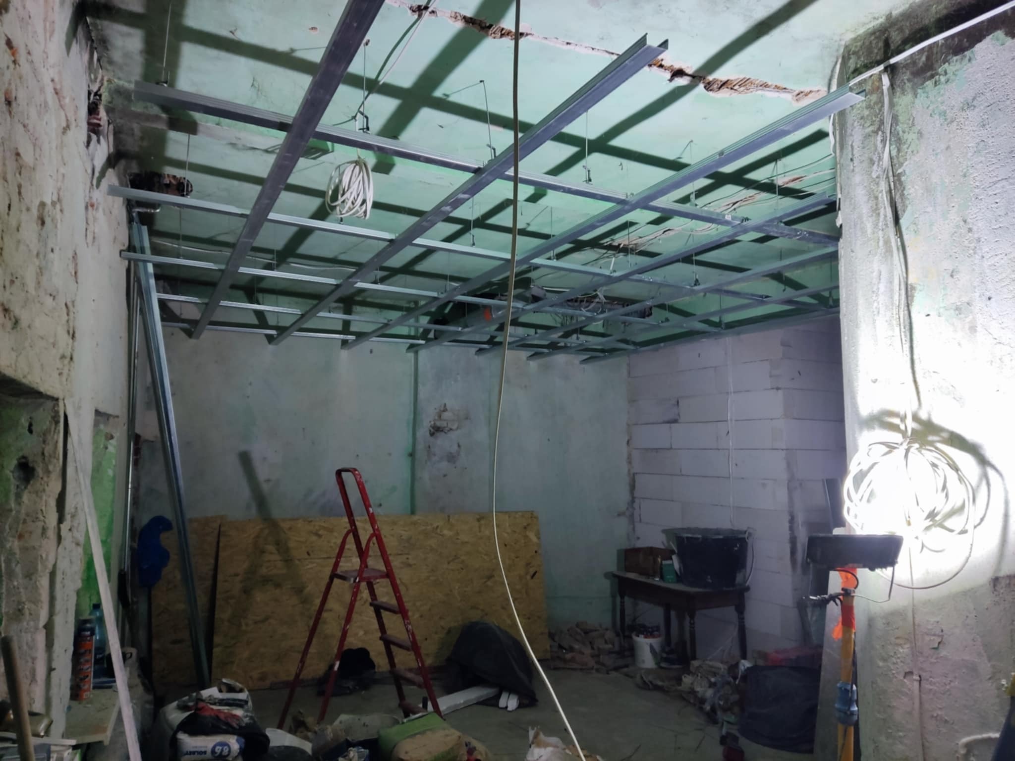 Montaż metalowej konstrukcji sufitu podwieszanego w surowym wnętrzu. Widoczna drabina, płyta OSB i narzędzia. Ściany z widocznymi śladami wcześniejszych prac.