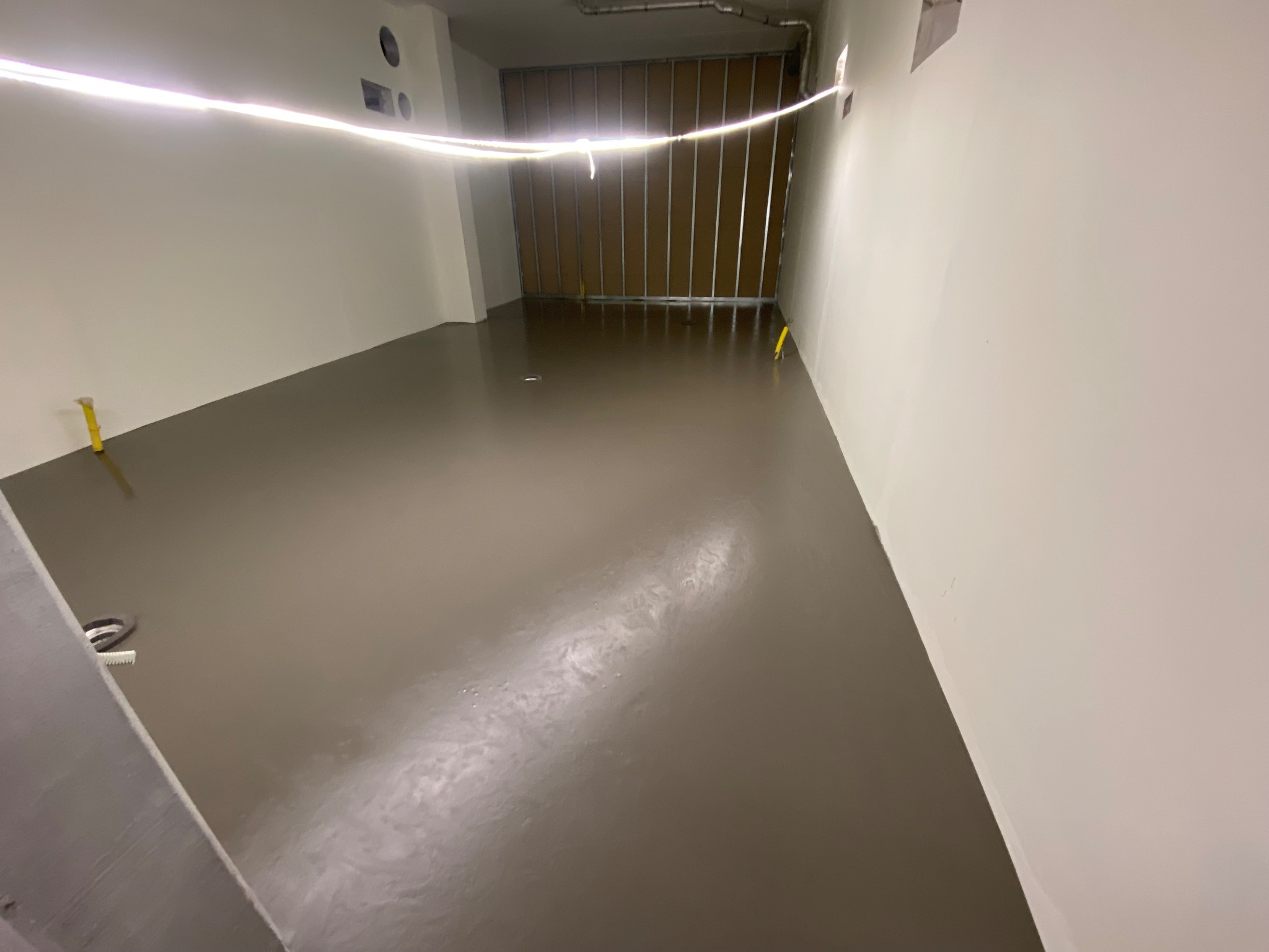 Gładka, brązowa wylewka samopoziomująca w jasnym pomieszczeniu, widoczne żółte znaczniki poziomu i metalowa konstrukcja ściany w tle.