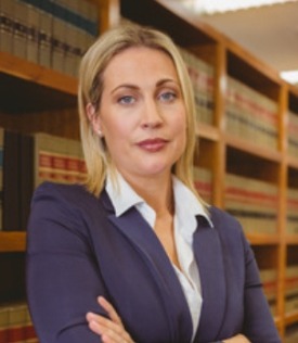 Portret kobiety w granatowym żakiecie, z blond włosami, stojącej na tle regałów z książkami prawniczymi.