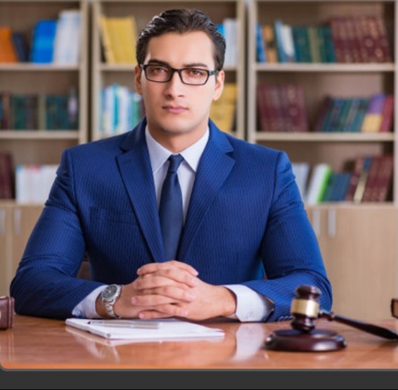 Portret adwokata w niebieskim garniturze siedzącego za biurkiem z drewnianym młotkiem sędziowskim i dokumentami na tle biblioteczki.