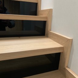 STOLARSTWO WOODSOL PIOTR SOLNICA - Detale jasnych, drewnianych schodów z czarnymi elementami, widoczne od dołu, z odbiciem osoby robiącej zdjęcie.