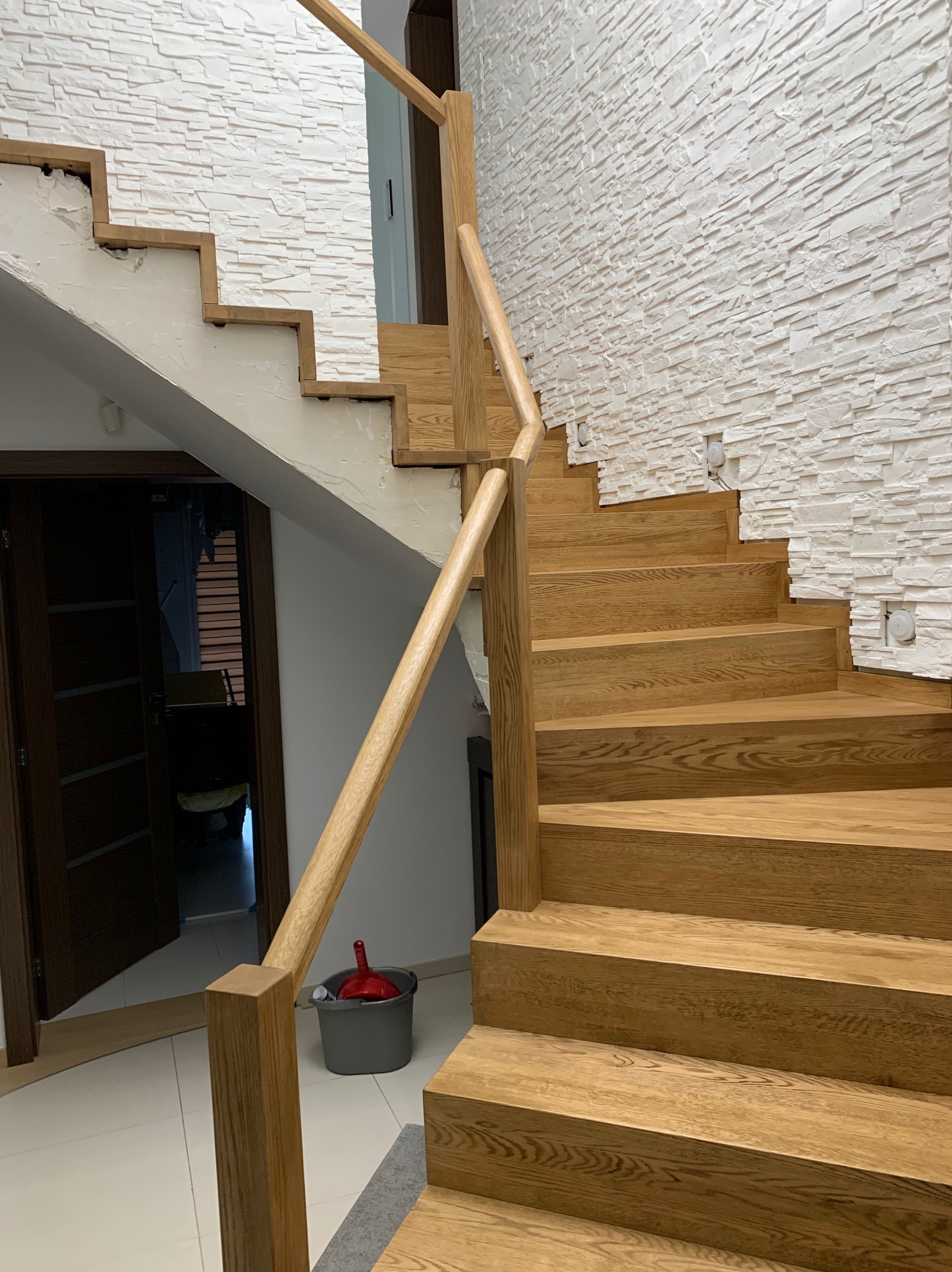 Jasne, drewniane schody z poręczą, prowadzące w górę wzdłuż ściany wykończonej dekoracyjnym kamieniem. Widoczny fragment drzwi i wiadro z akcesoriami malarskimi.