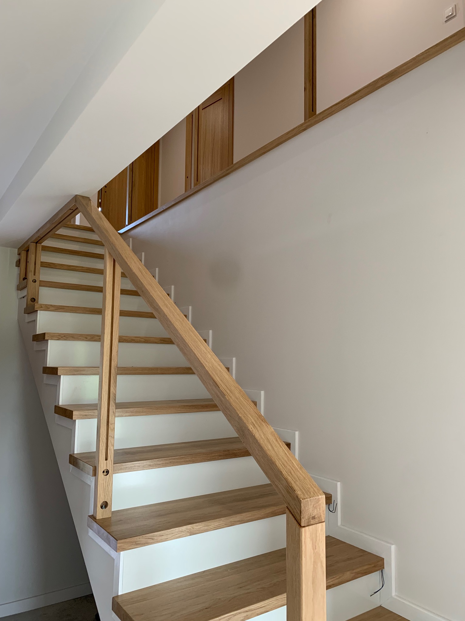 Jasne, drewniane schody z białymi podstopnicami i prostą drewnianą balustradą w nowoczesnym wnętrzu. Widoczne mocowania balustrady do stopni.