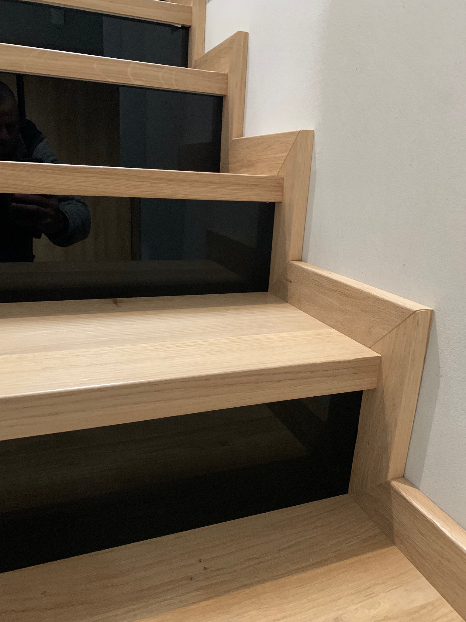 Detale jasnych, drewnianych schodów z czarnymi elementami, widoczne od dołu, z odbiciem osoby robiącej zdjęcie.