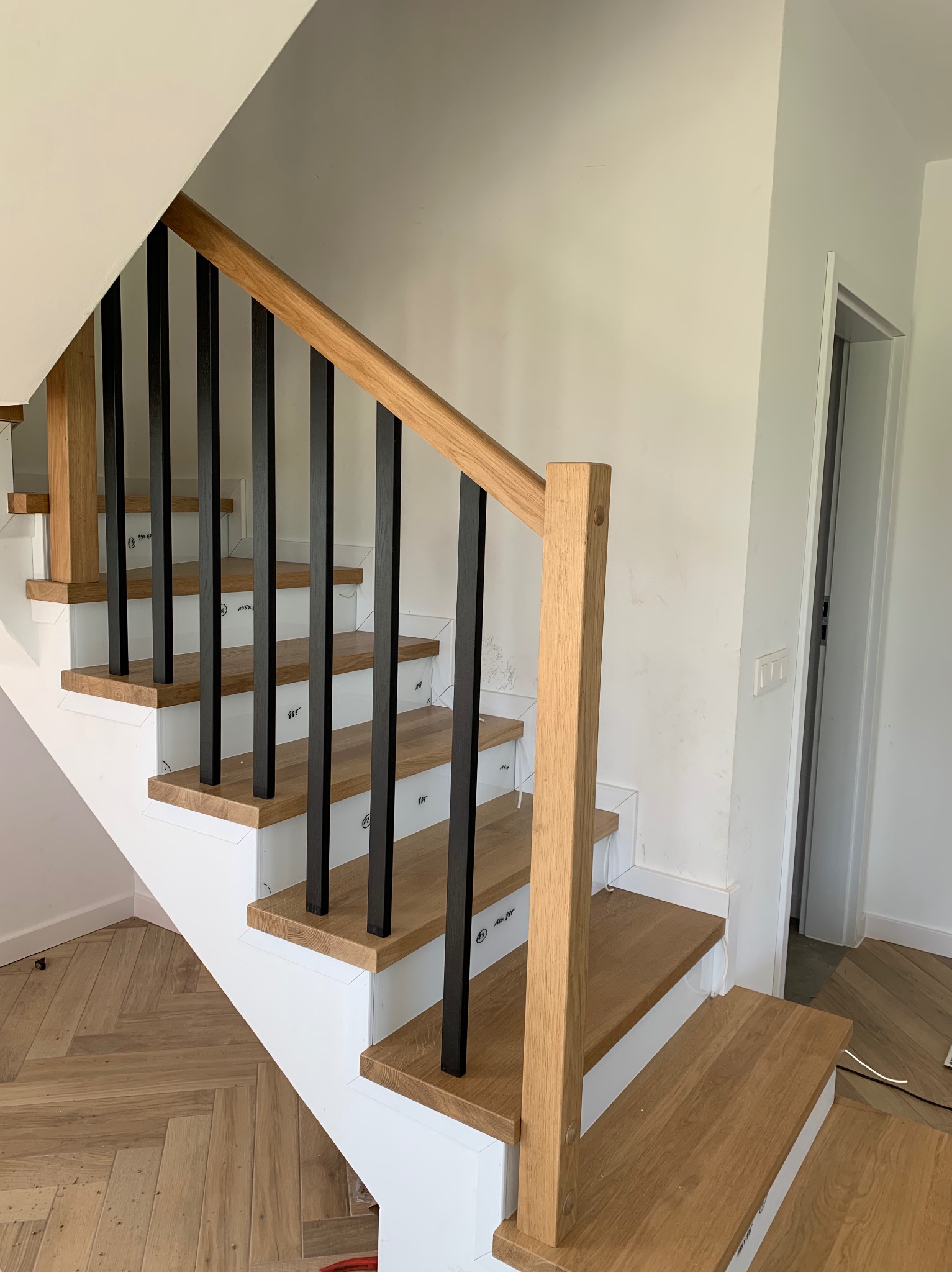 Nowoczesne schody z drewnianymi stopniami i czarnymi, kwadratowymi balustradami, częściowo zabezpieczone folią ochronną, widoczne ślady prac wykończeniowych na ścianie.