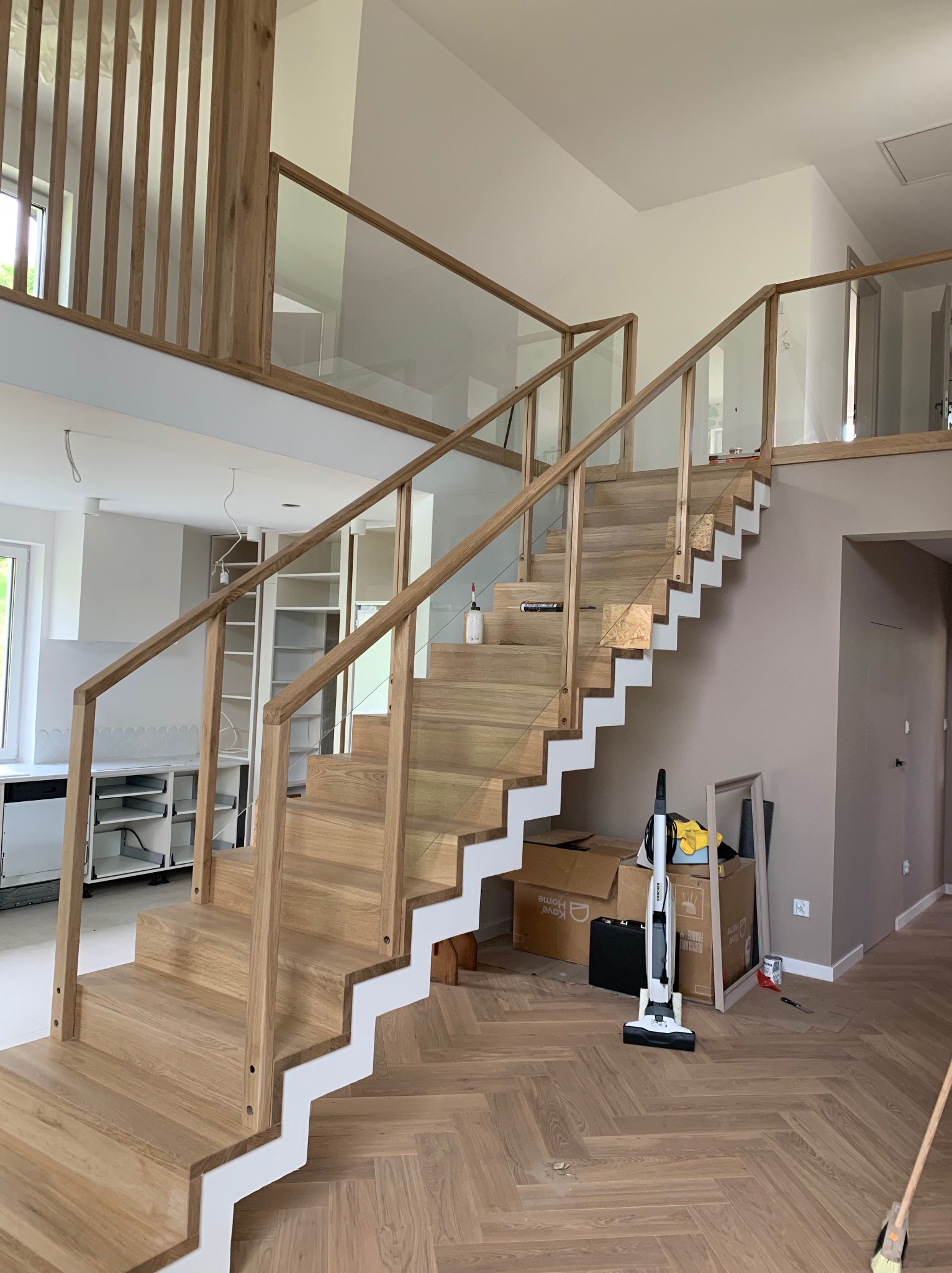 Jasne drewniane schody z białymi podstopnicami i szklaną balustradą w dwupoziomowym domu, widoczne narzędzia i kartony na podłodze.