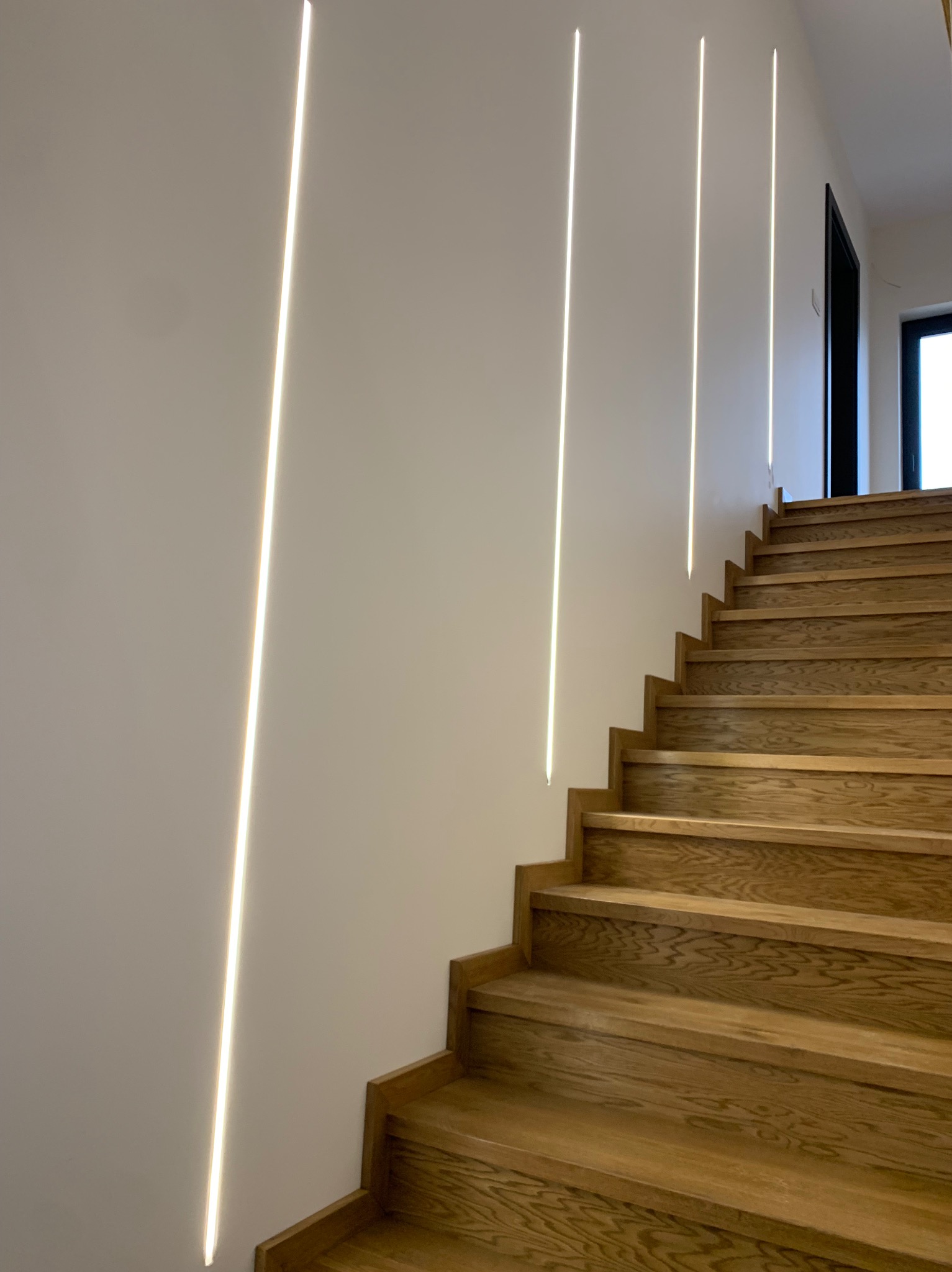 Nowoczesne schody z drewna o naturalnym odcieniu, oświetlone pionowymi paskami LED w ścianie, widok z dołu.
