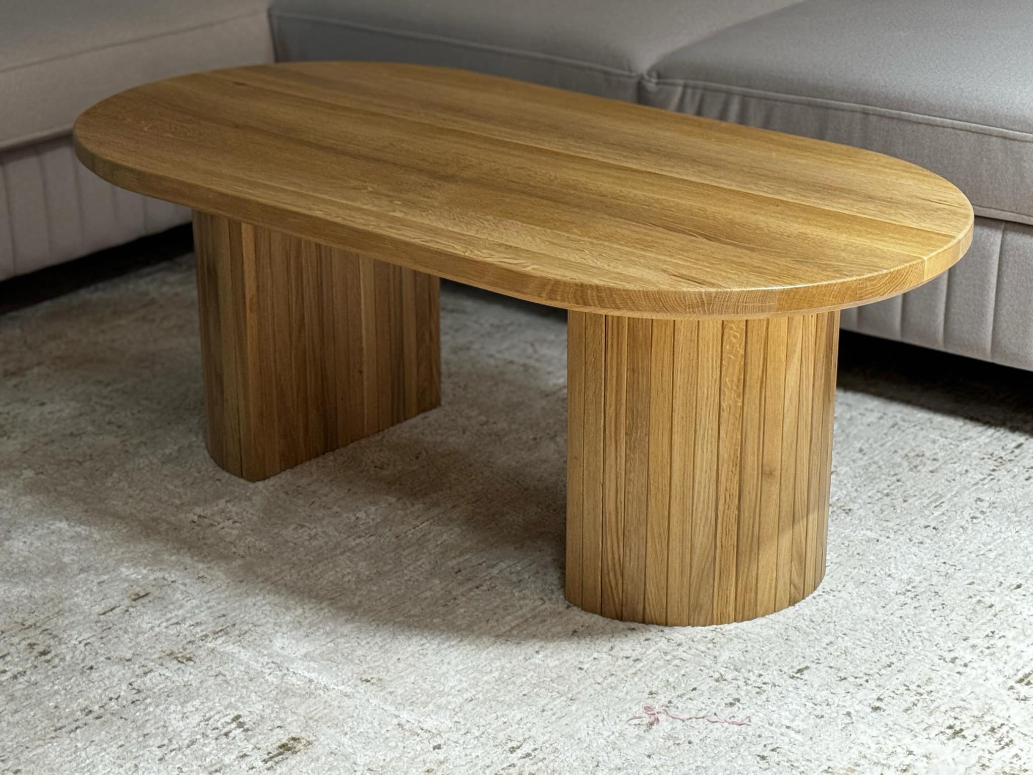Owalny stolik kawowy z jasnego drewna z charakterystycznymi, żłobionymi nogami ustawiony na wzorzystym dywanie, obok szarej sofy.