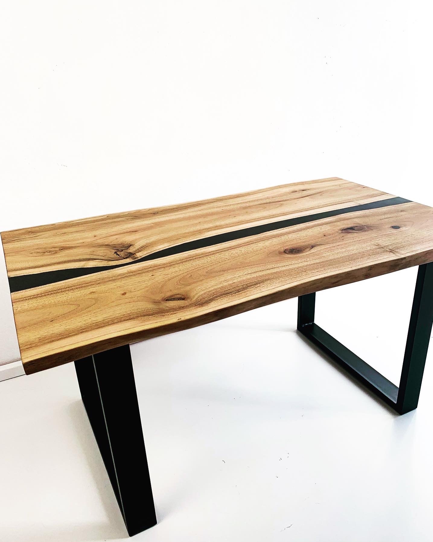 Blat z jasnego drewna z żywicą epoksydową w kolorze czarnym, osadzony na metalowych, czarnych nogach w kształcie trapezu i prostokąta, na białym tle.