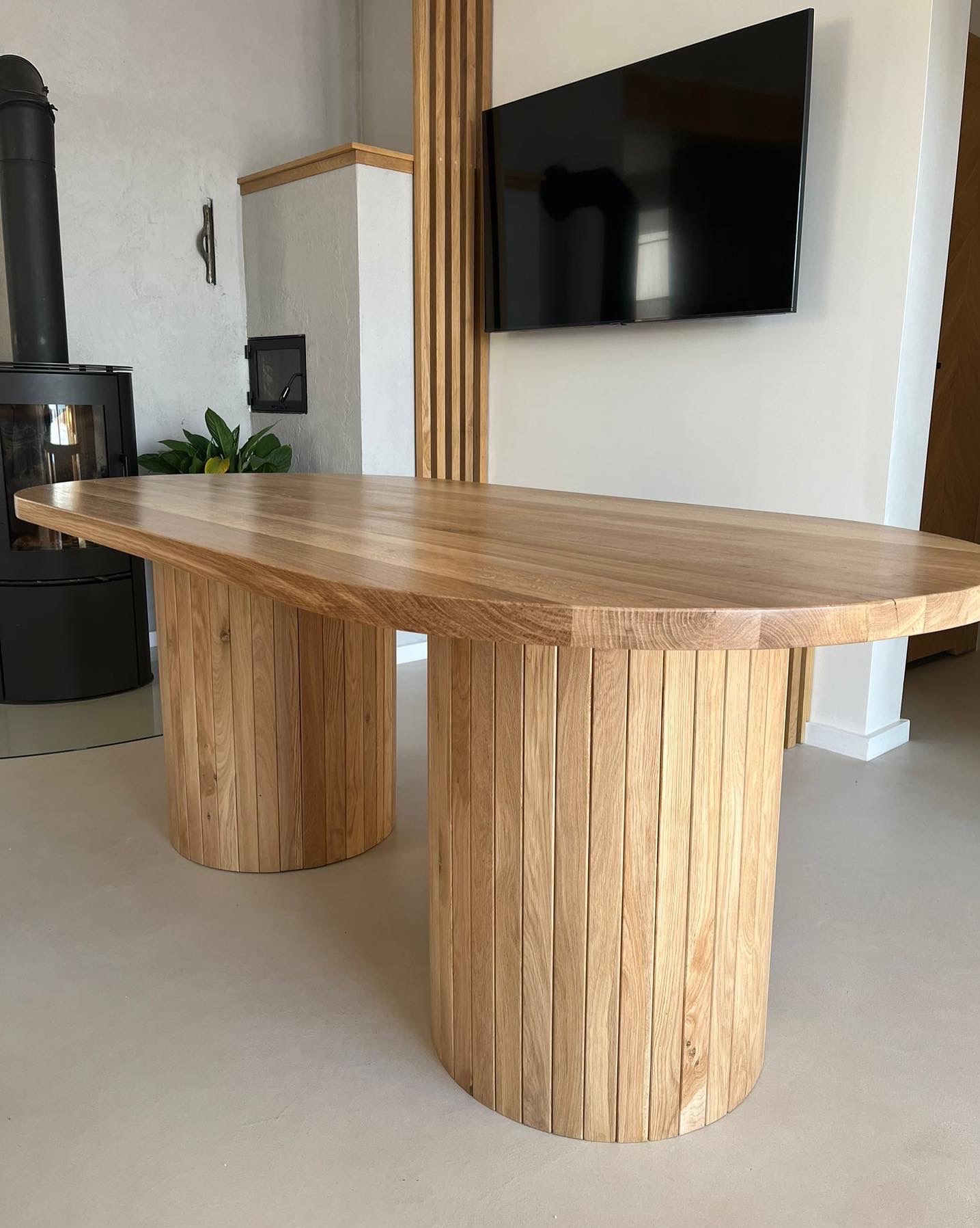 Owalny stół z blatem z jasnego drewna i cylindrycznymi nogami z pionowych listewek drewnianych, na tle minimalistycznego wnętrza z kominkiem i telewizorem.