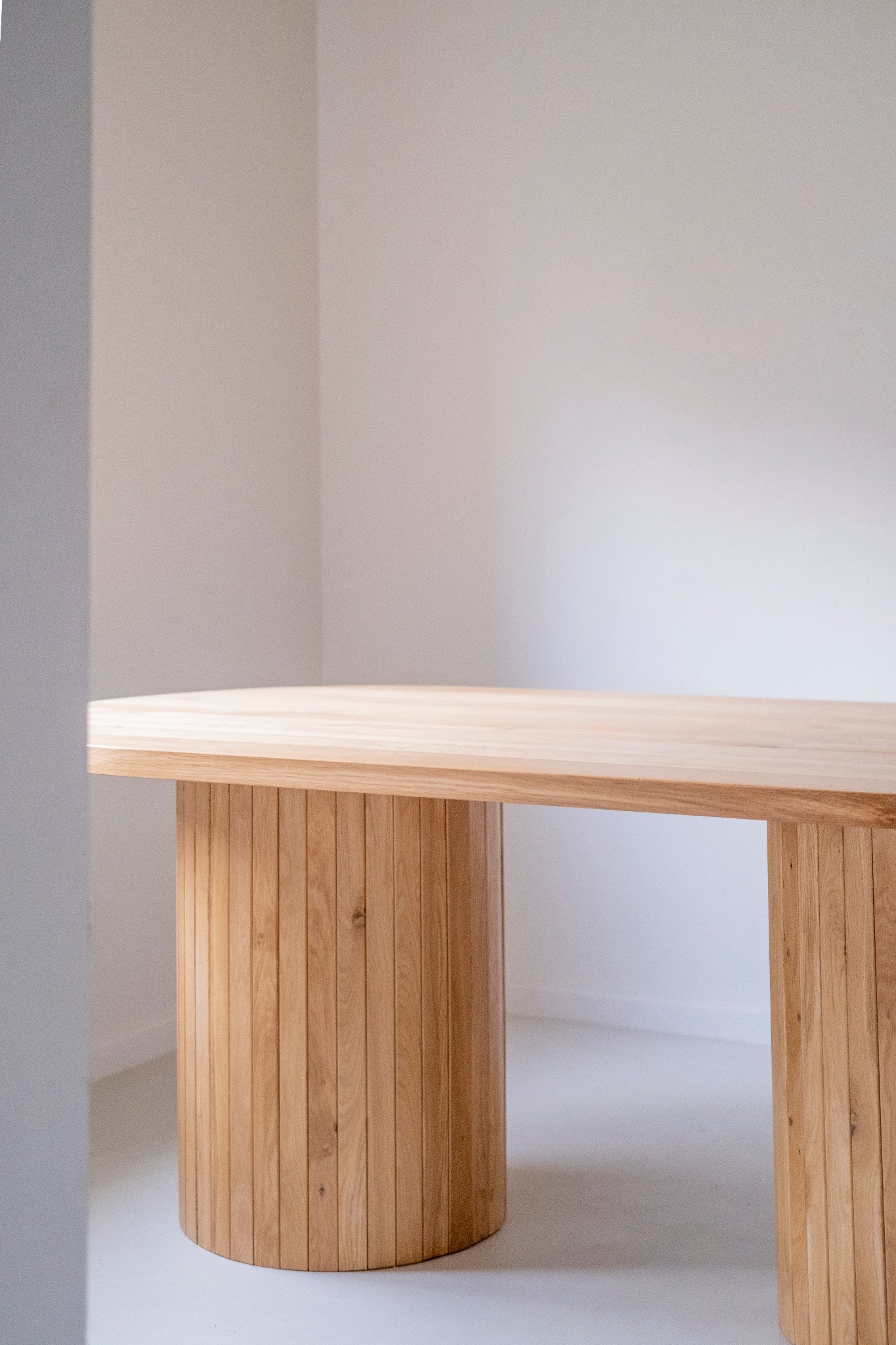 Jasny, minimalistyczny blat z drewna o zaokrąglonym kształcie, wsparty na dwóch cylindrycznych nogach z pionowych listew, na tle białej ściany i podłogi.