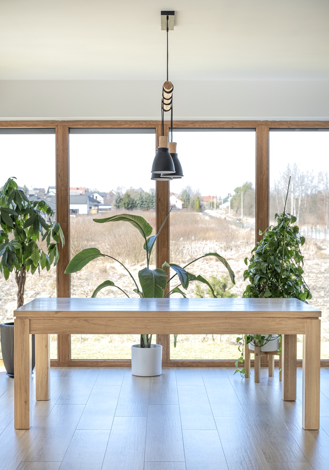 Jasny, drewniany blat stołu w minimalistycznym wnętrzu z dużym oknem, roślinami doniczkowymi i nowoczesną lampą wiszącą.