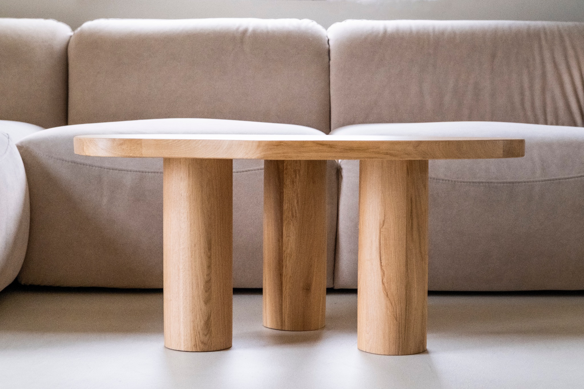 Minimalistyczny stolik kawowy z jasnego drewna z trzema cylindrycznymi nogami, umieszczony przed beżową sofą modułową, ukazujący prostotę i naturalne materiały.