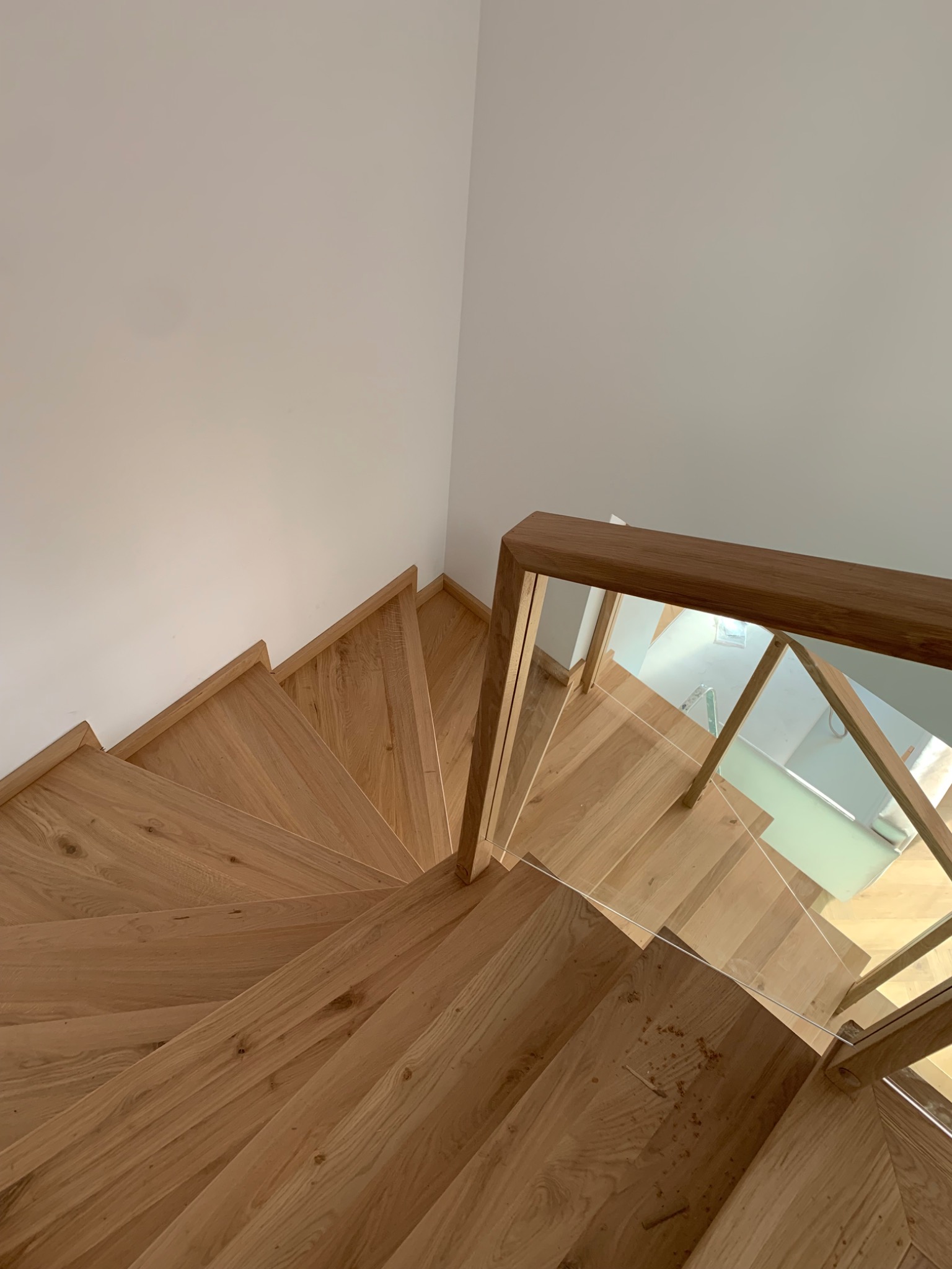 Spiralne schody drewniane z jasnego drewna dębowego, zabezpieczone szklaną balustradą, widok z góry na zakręcające stopnie.