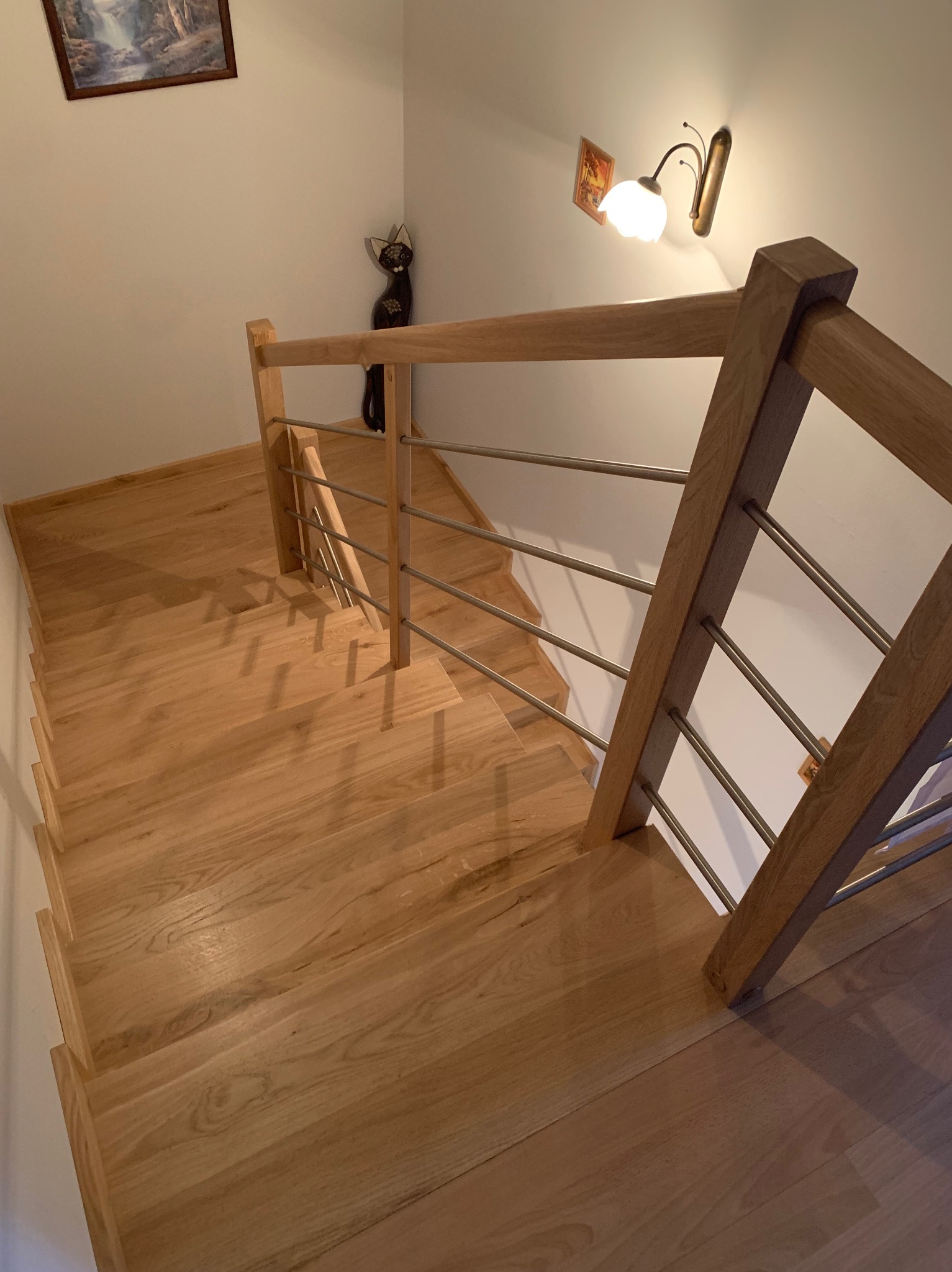 Widok z góry na jasne, drewniane schody z metalowymi balustradami, oświetlone lampą ścienną, z dekoracyjną figurką kota w tle.