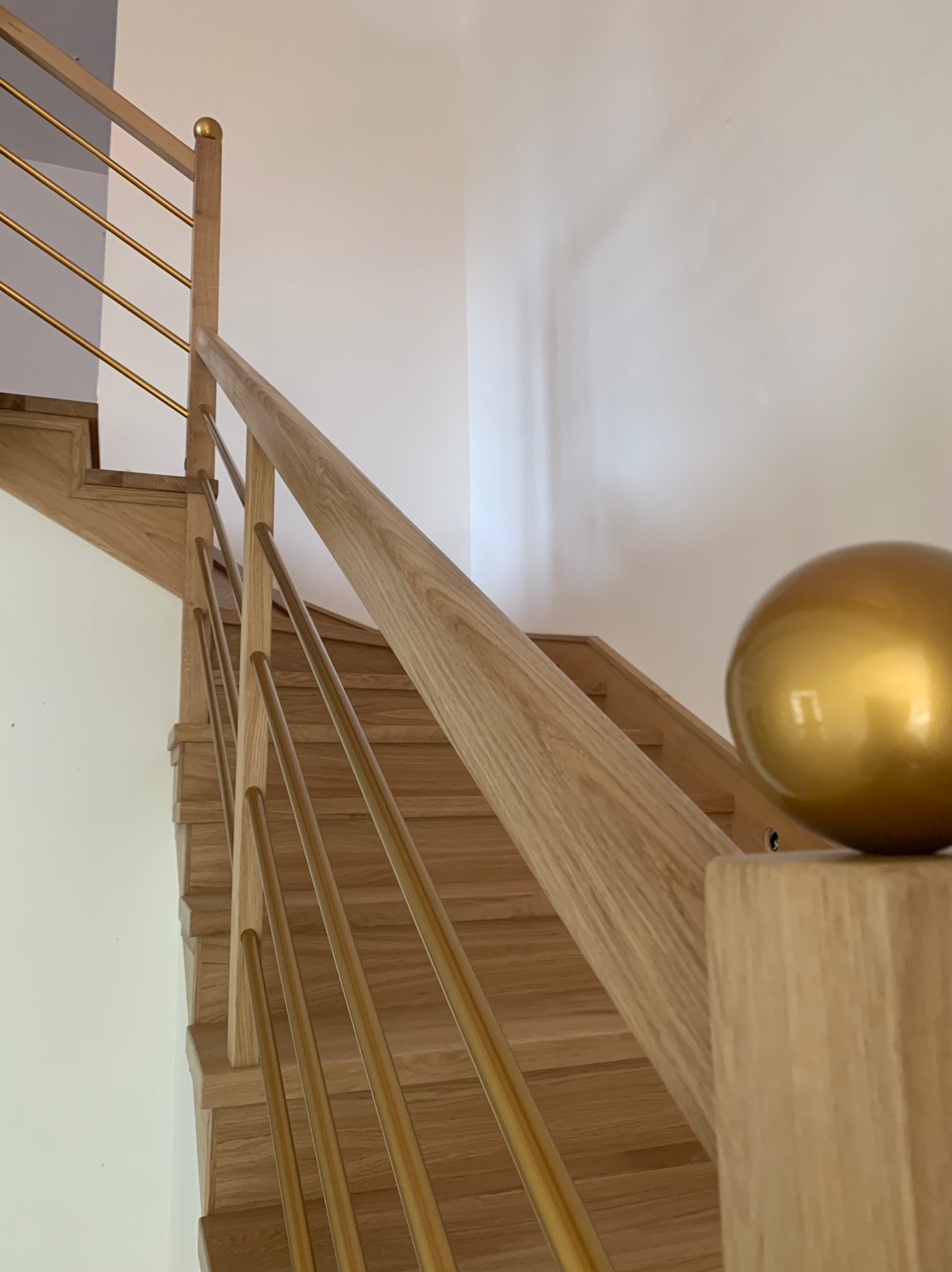 Jasne drewniane schody z metalowymi, złotymi balustradami i dekoracyjną złotą kulą na słupku.
