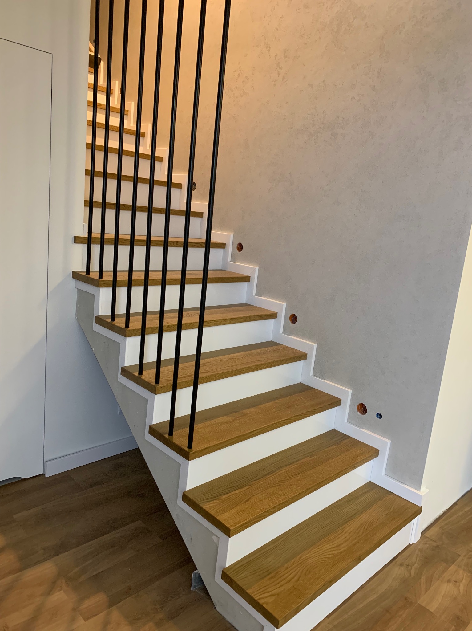 Nowoczesne schody z drewnianymi stopniami, białymi podstopnicami i czarną metalową balustradą, widok z dołu, ściana z dekoracyjnym tynkiem.