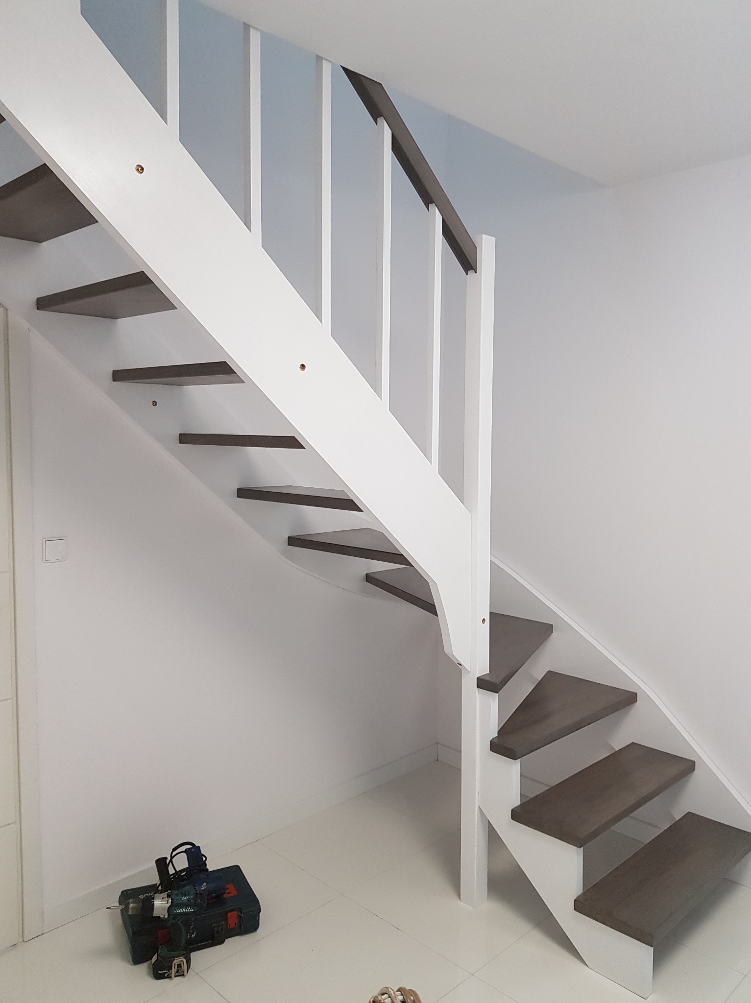 Białe schody z drewnianymi stopniami i balustradą, widoczne narzędzia montażowe w rogu pomieszczenia, minimalistyczny design.