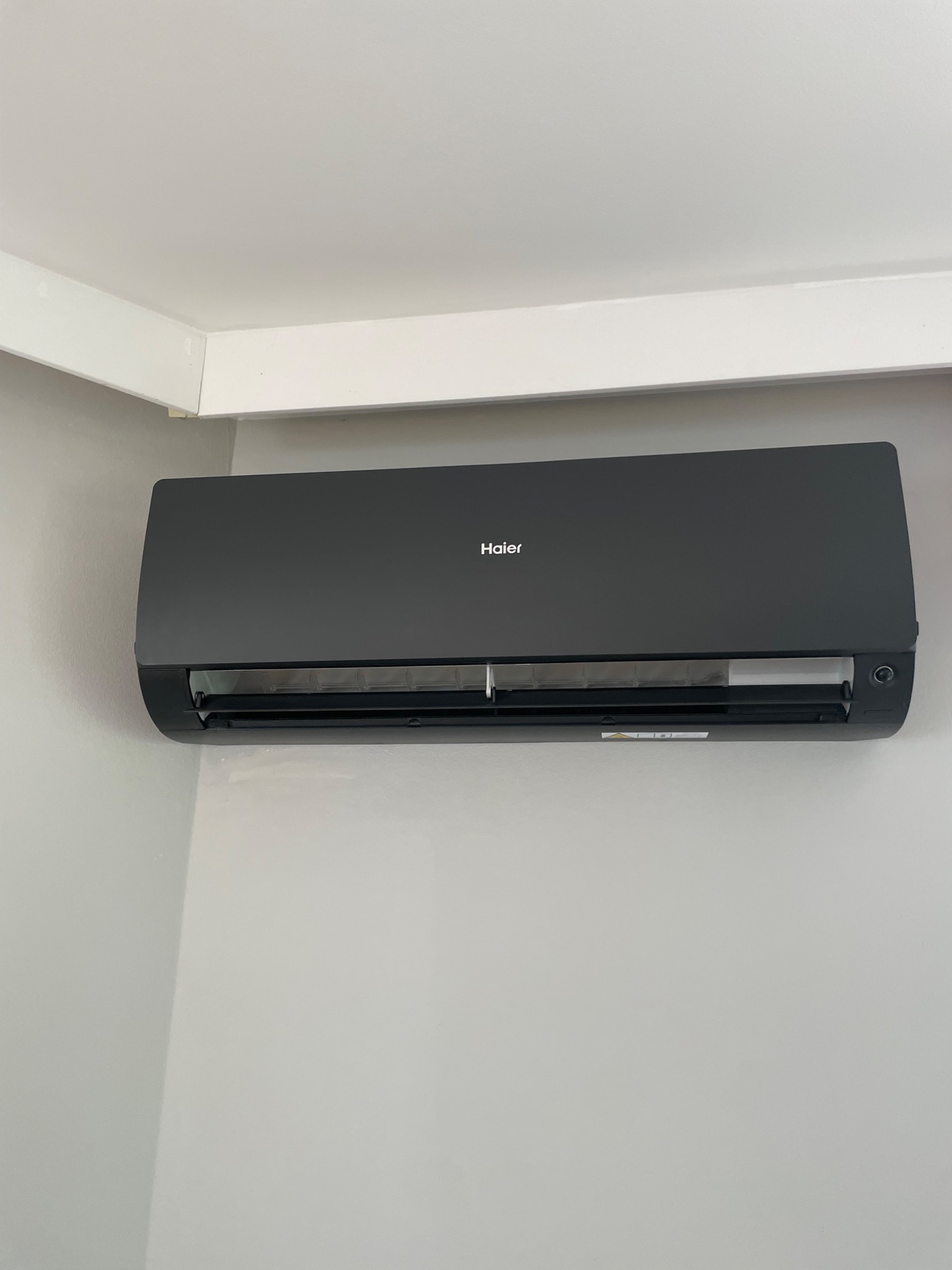 Czarny klimatyzator Haier zamontowany na szarej ścianie w pomieszczeniu z białym sufitem i listwą. Widok z dołu, centralnie.