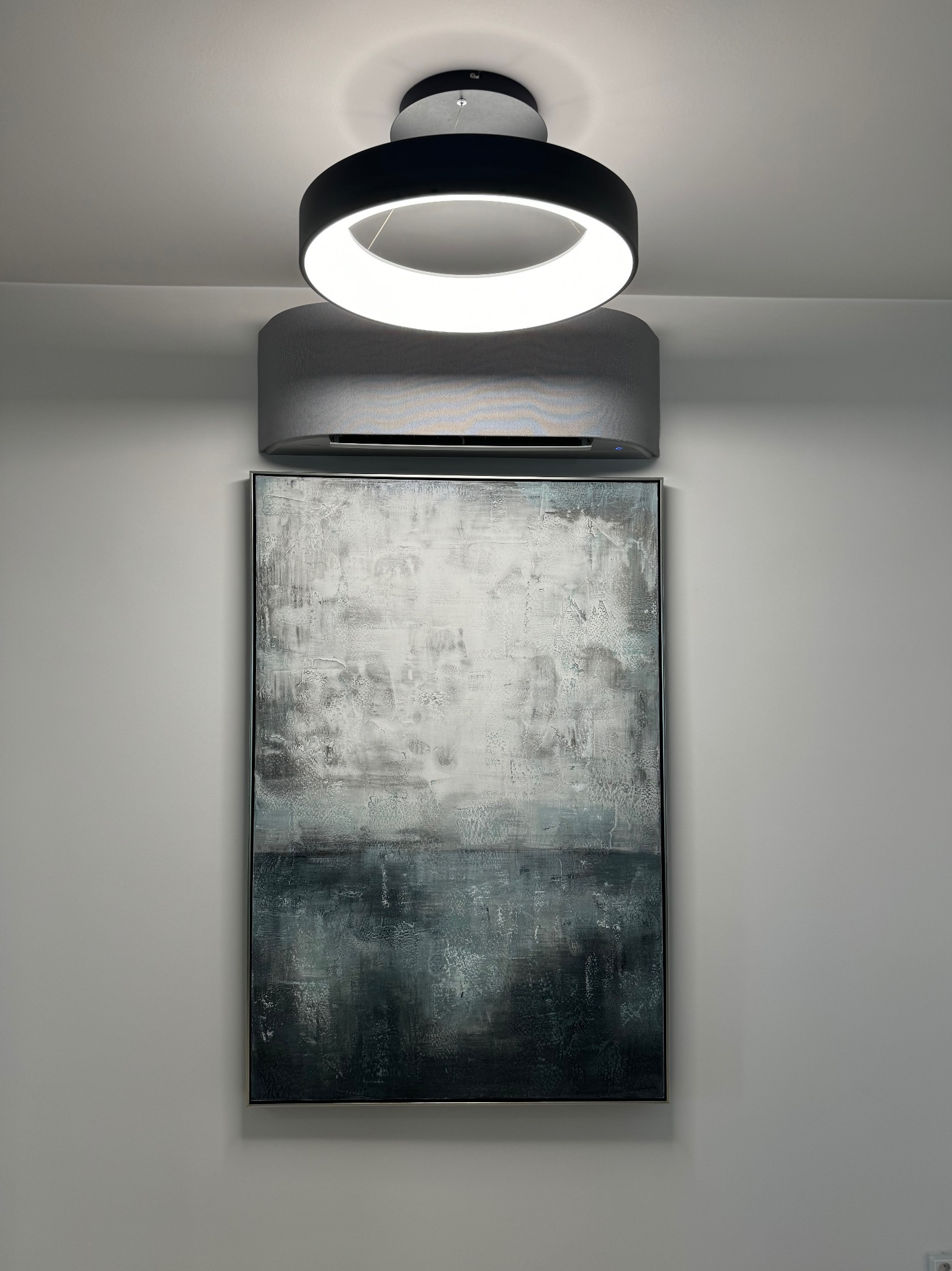 Szara klimatyzacja ścienna w nowoczesnym wnętrzu, z okrągłą lampą sufitową i abstrakcyjnym obrazem w ramie. Minimalistyczny design, stonowana kolorystyka.