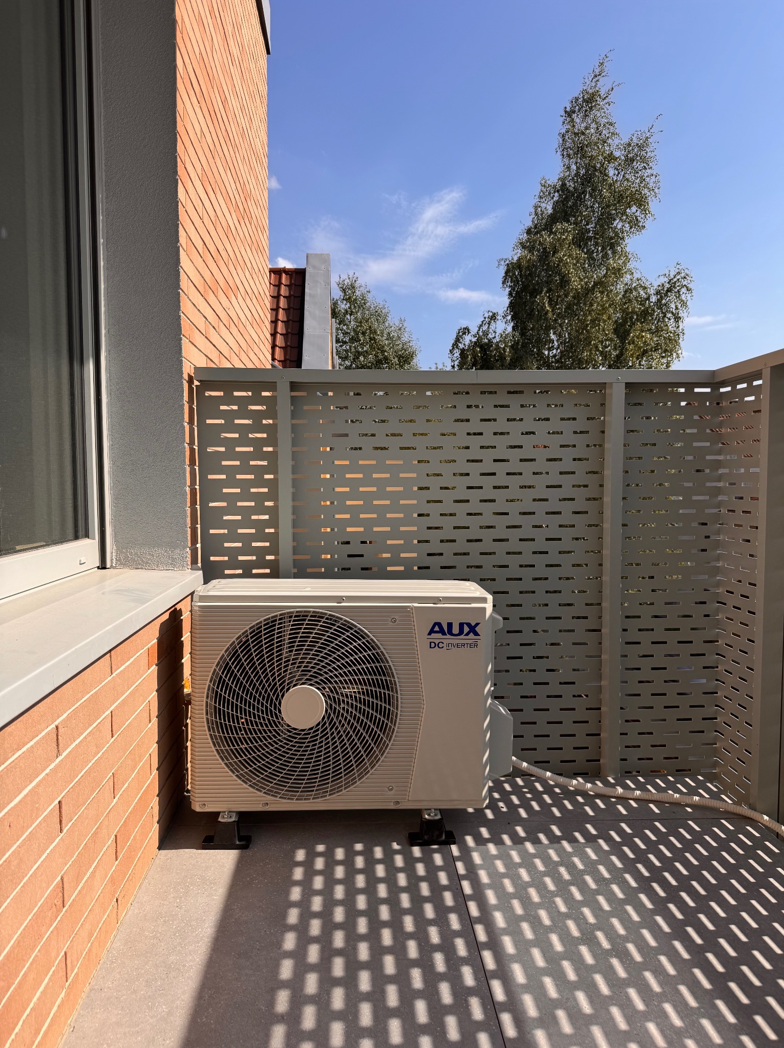 Biała jednostka zewnętrzna klimatyzacji AUX DC Inverter zamontowana na balkonie. Cień ażurowej osłony na podłodze tworzy wzór. Widok na błękitne niebo.