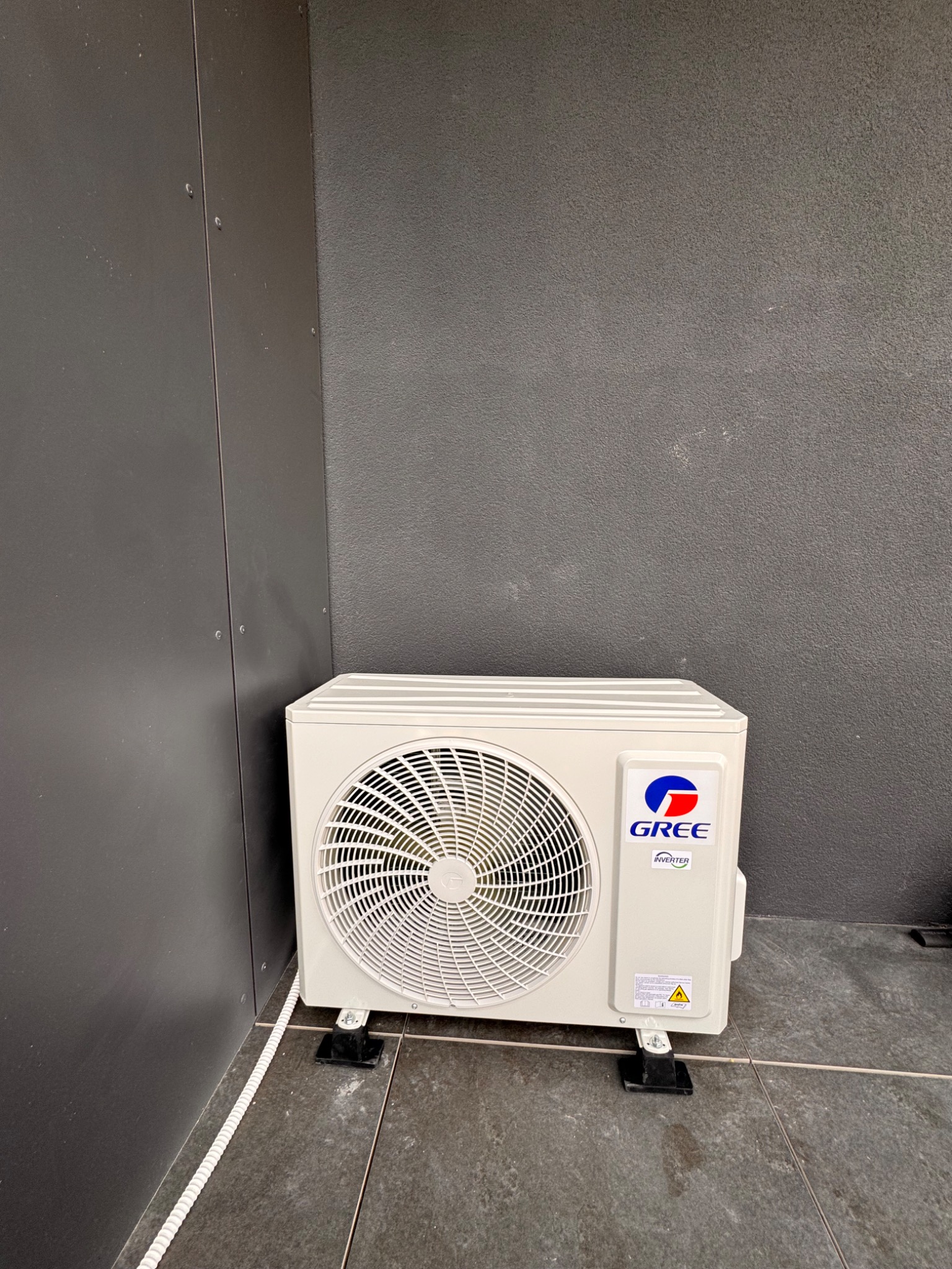 Zewnętrzna jednostka klimatyzacji GREE na grafitowej elewacji z widoczną instalacją elektryczną. Jednostka stoi na czarnych wspornikach na szarych płytkach.