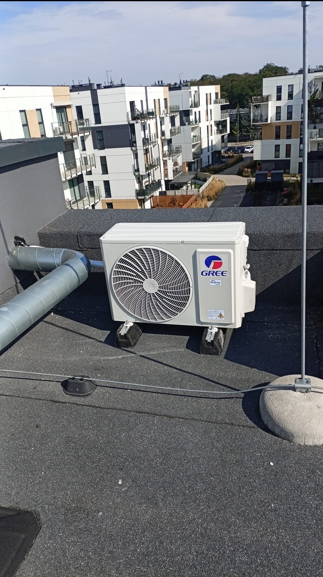 Biała jednostka zewnętrzna klimatyzacji Gree na dachu bloku mieszkalnego, widoczne rury i osprzęt montażowy. Na drugim planie budynki.