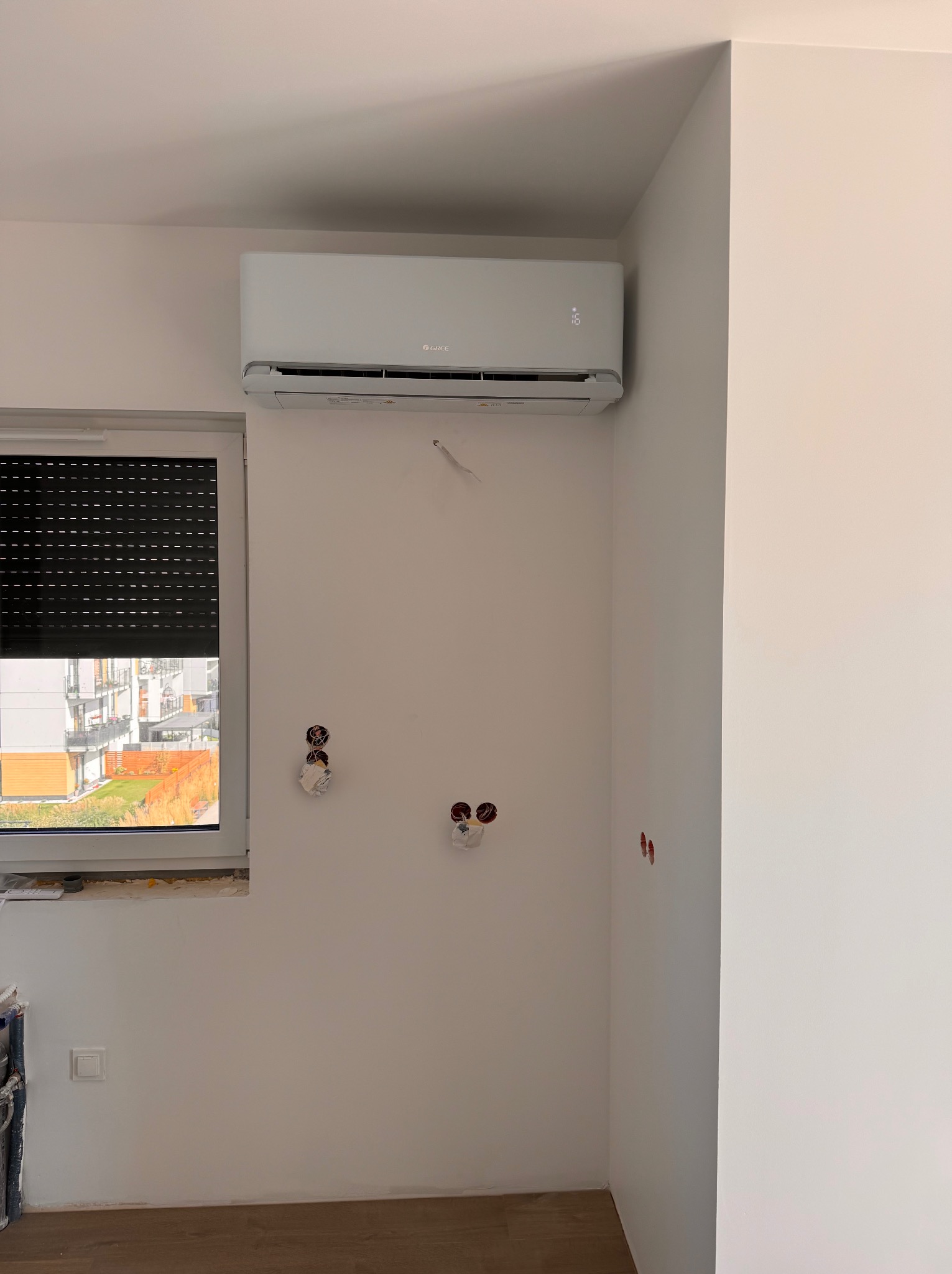 Biały klimatyzator Gree zamontowany na ścianie w jasnym pokoju, widoczne przygotowane punkty elektryczne i okno z roletą zewnętrzną.