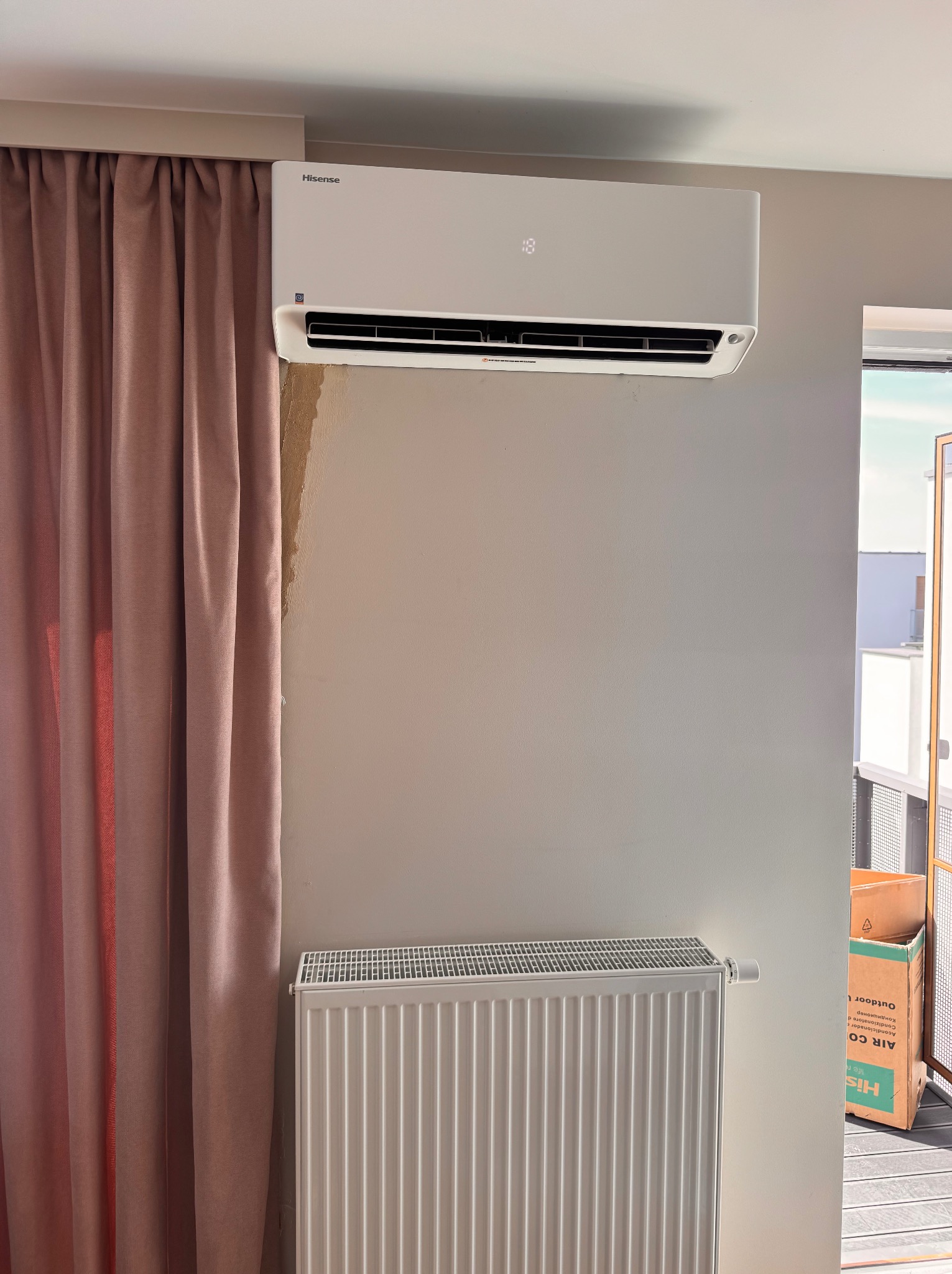 Biały klimatyzator Hisense na ścianie w pokoju z beżowymi zasłonami i grzejnikiem. Widoczne ślady po montażu. Na balkonie karton z napisem AIR CO.