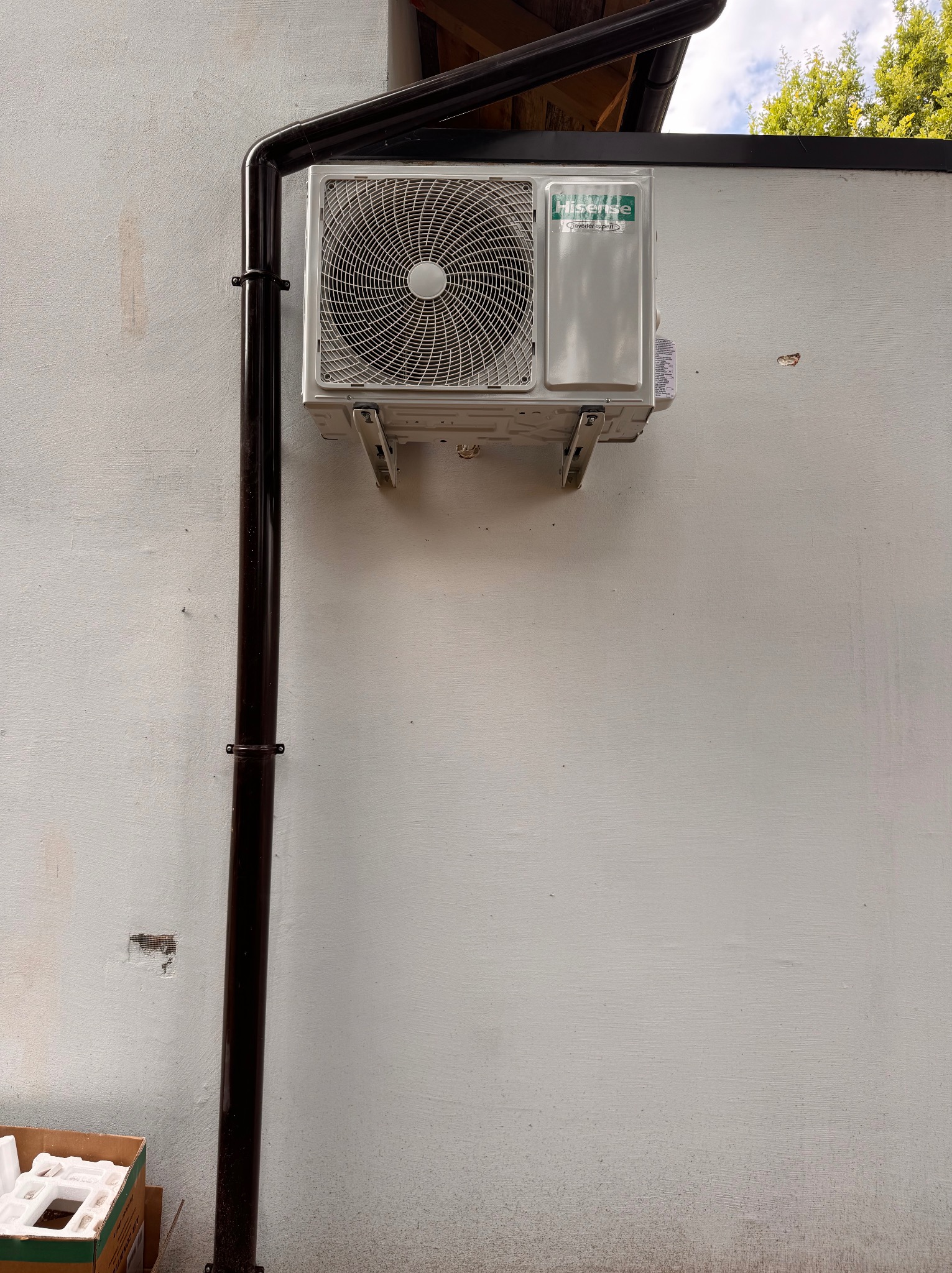 Biała klimatyzacja Hisense zamontowana na jasnej ścianie zewnętrznej budynku obok brązowej rynny spustowej. Widoczny fragment kartonu pod klimatyzatorem.