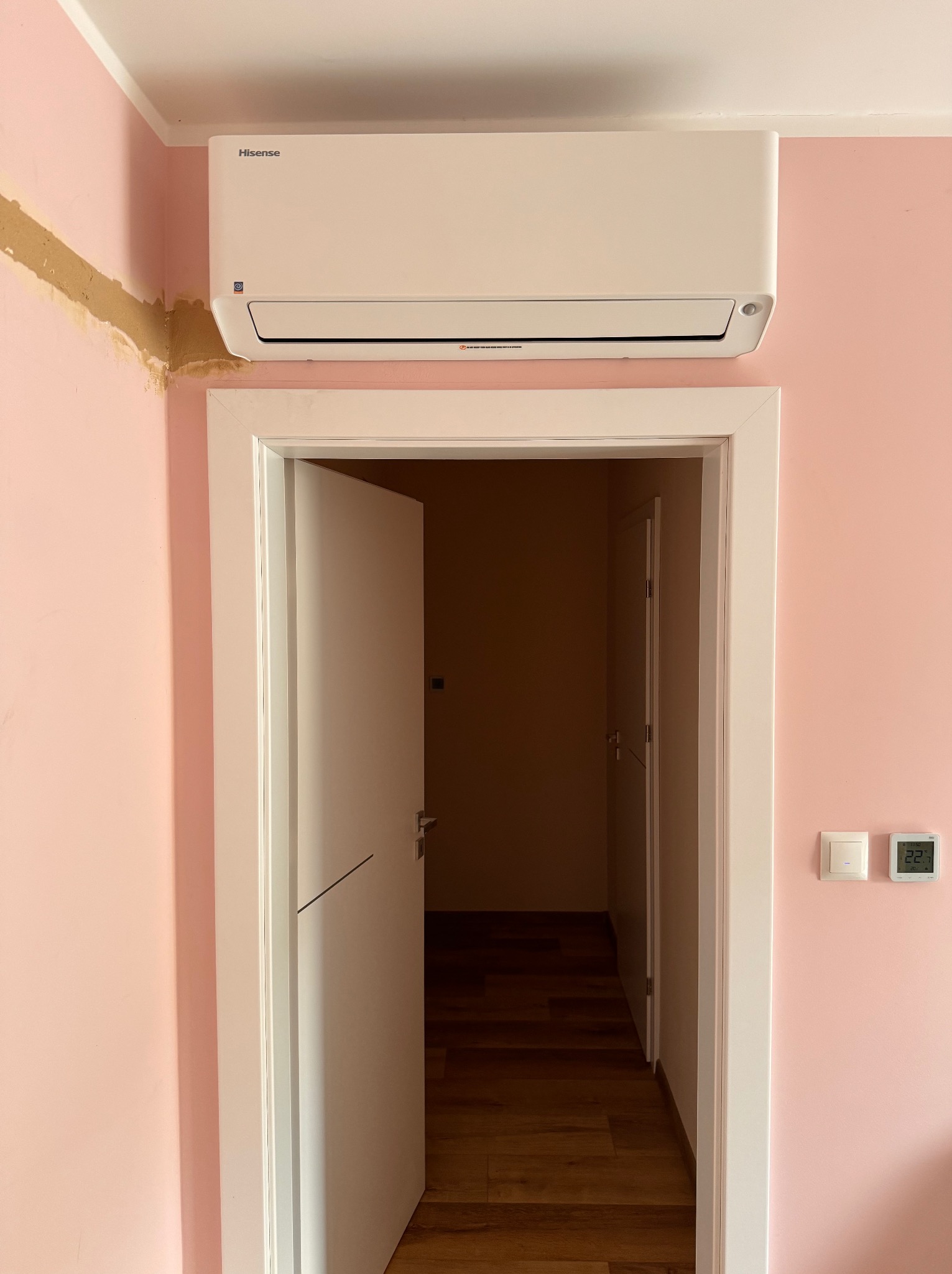 Biała klimatyzacja Hisense na różowej ścianie nad otwartymi drzwiami. Widoczny panel sterowania i fragment uszkodzonej ściany. W tle parkiet.