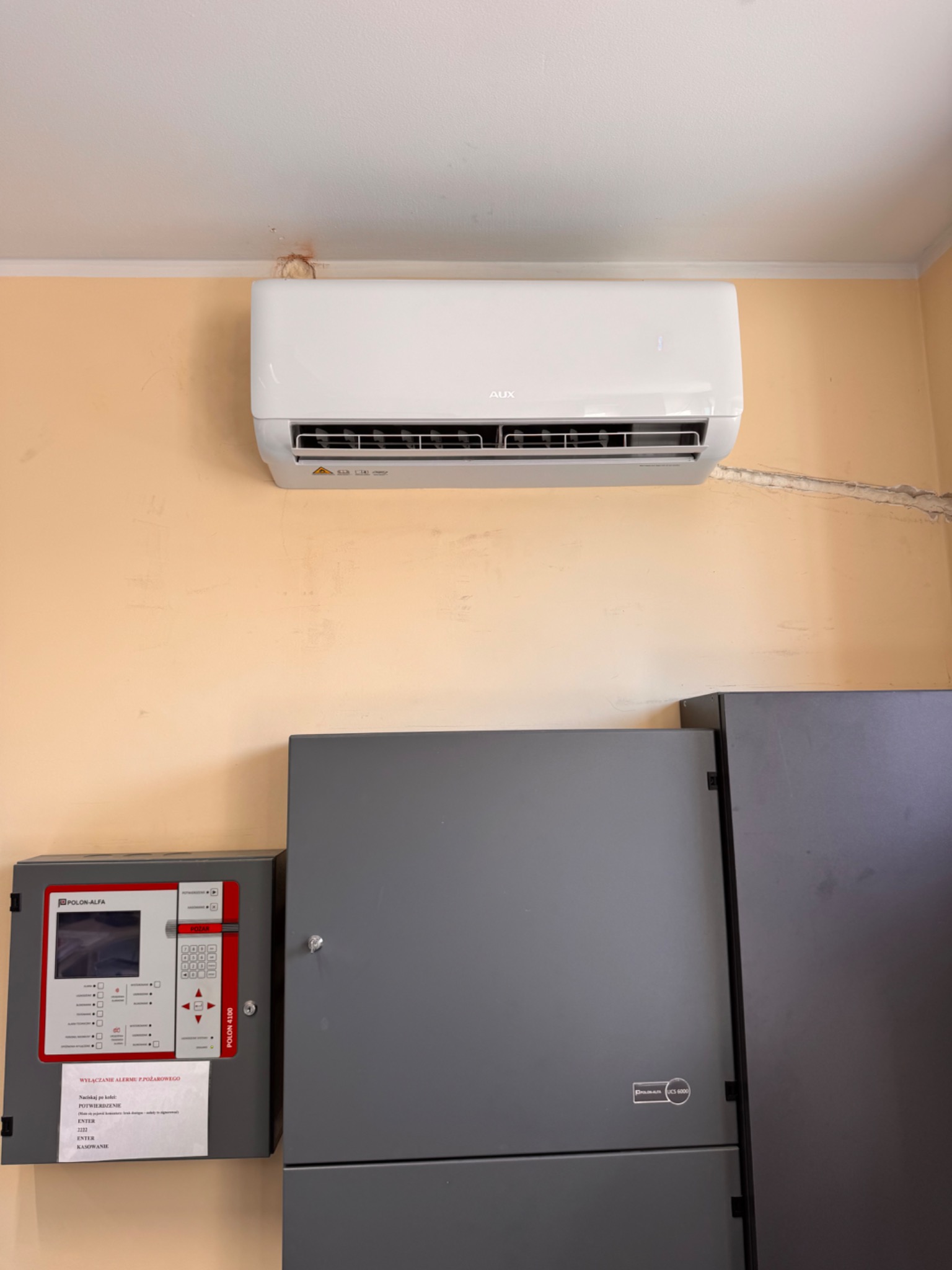 Biały klimatyzator AUX zamontowany na beżowej ścianie nad szarą szafką z panelem alarmowym Polon-Alfa. Widoczny fragment sufitu z plamą.