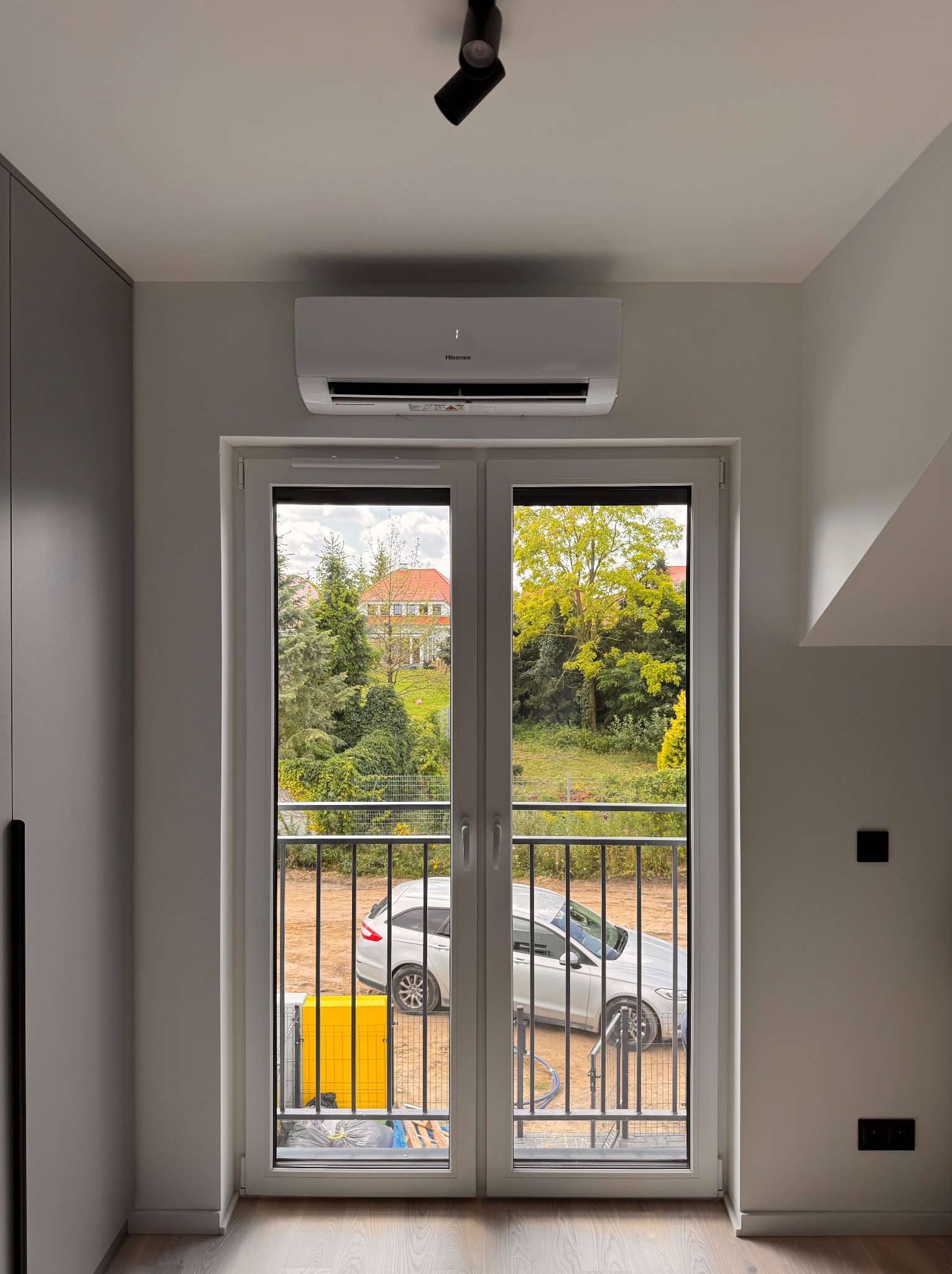 Biały klimatyzator Hisense nad balkonem w nowoczesnym wnętrzu. Na balkonie widoczny żółty agregat i samochód. Za balkonem zieleń drzew i dom.