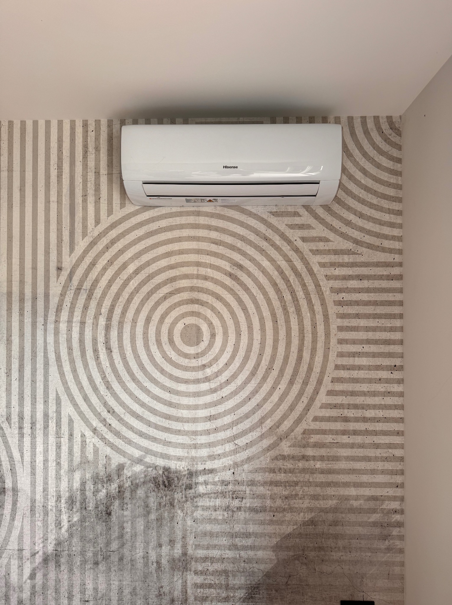 Biała klimatyzacja Hisense zamontowana na ścianie z geometrycznym wzorem w odcieniach szarości, widok z dołu, minimalistyczny design wnętrza.