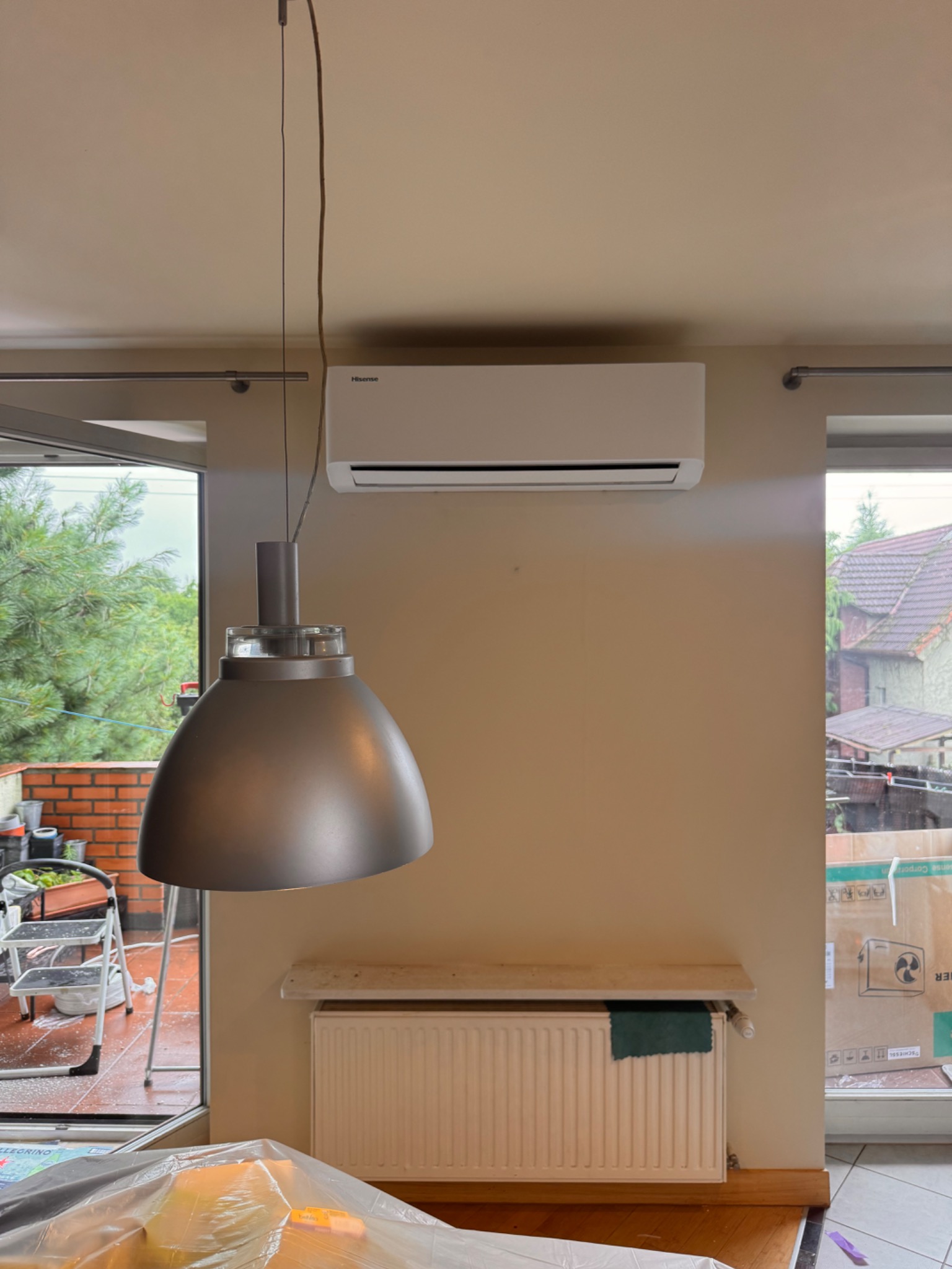 Zamontowany klimatyzator Hisense nad grzejnikiem w jasnym wnętrzu. Widoczny fragment lampy i okna z zielenią. Kartonowe pudła w tle.