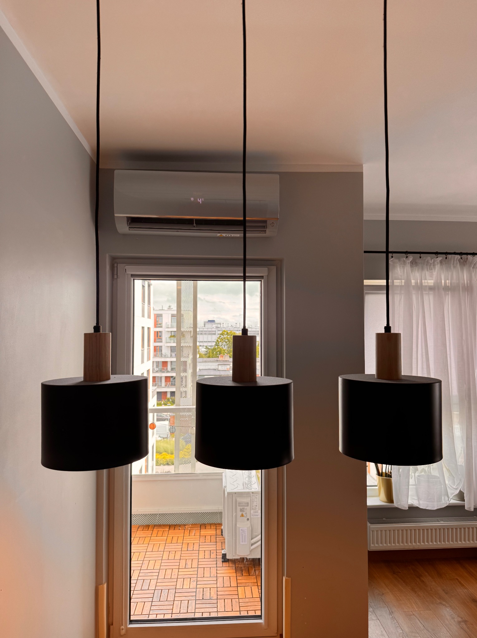 Biała klimatyzacja Haier nad balkonem z terakotą, widoczna przez trzy czarne lampy wiszące w szarym pokoju z białymi zasłonami i grzejnikiem.