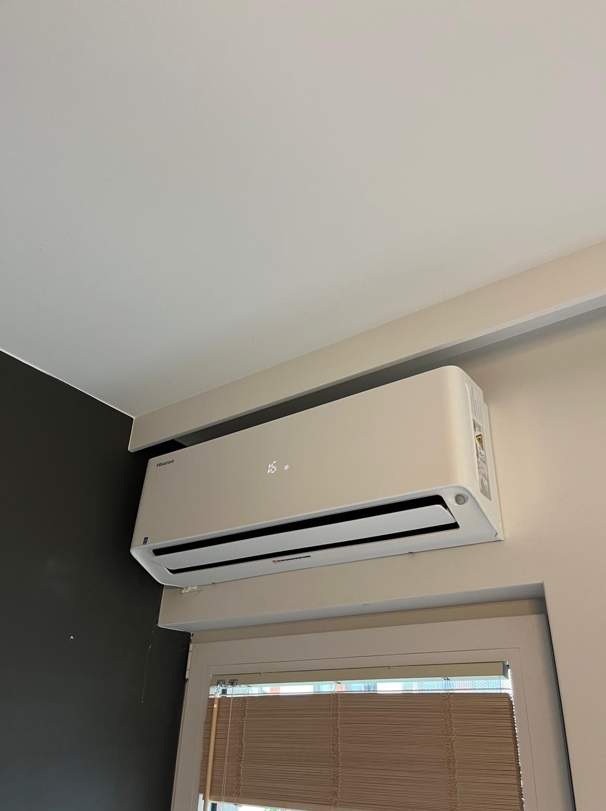 Biały klimatyzator Hisense zamontowany na ścianie nad oknem z żaluzjami, wyświetlający temperaturę 16 stopni, na tle ciemnej i jasnej ściany.