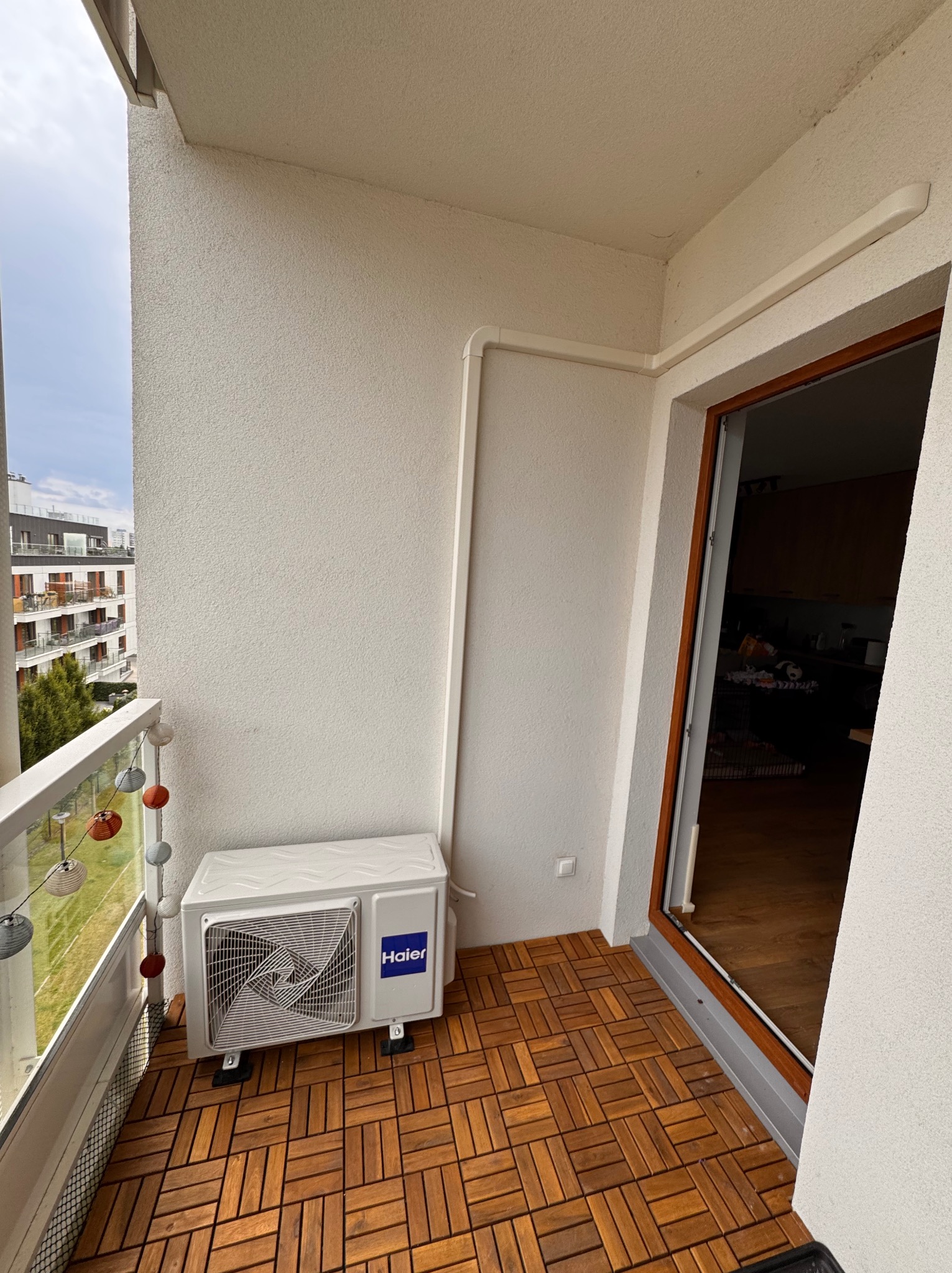 Biała jednostka zewnętrzna klimatyzacji Haier zamontowana na drewnianej podłodze balkonu z widokiem na miasto.