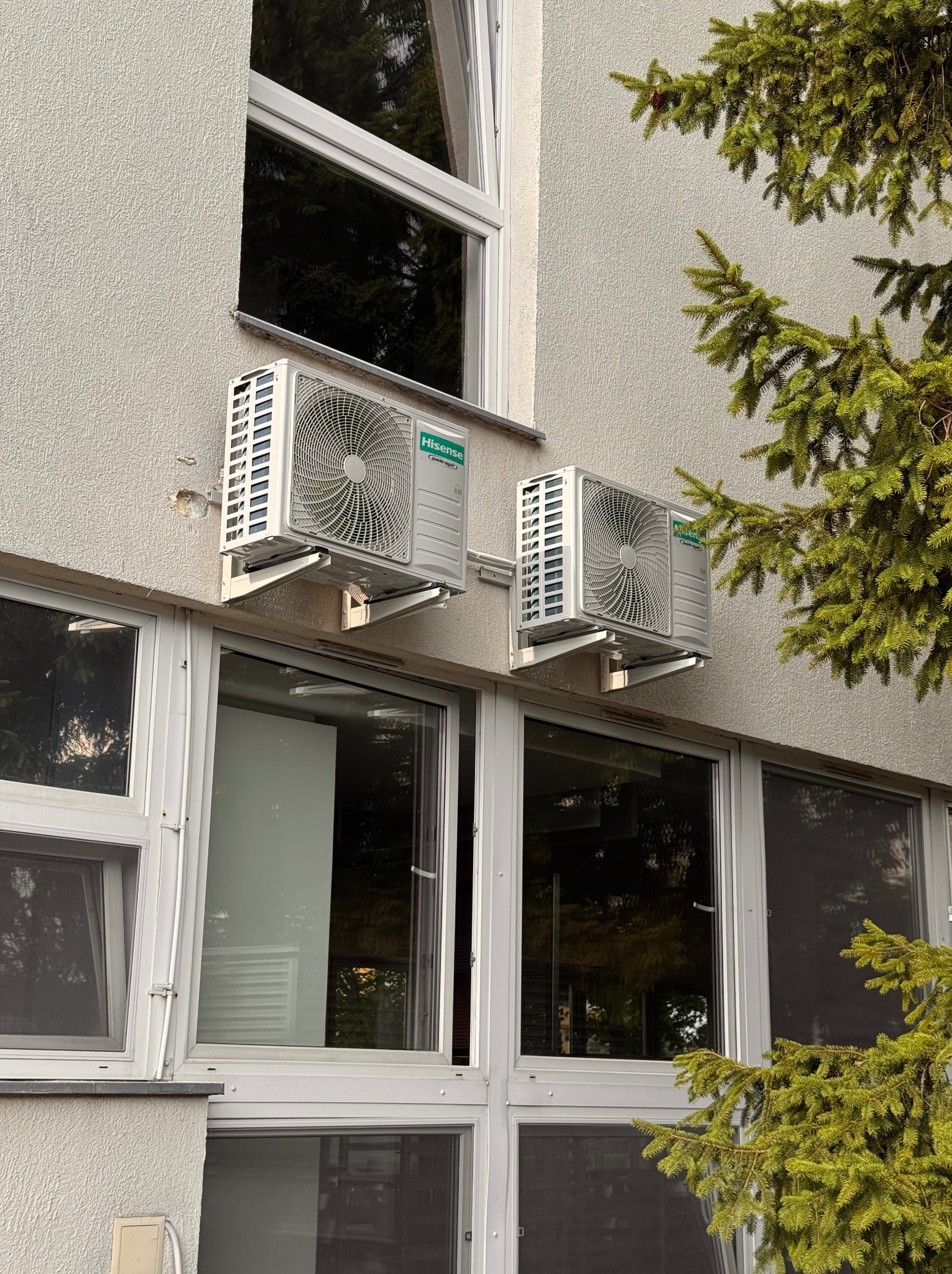 Dwa klimatyzatory Hisense zamontowane na elewacji budynku, widoczne z zewnątrz, z fragmentem drzewa iglastego po prawej stronie. Jasna elewacja i okna.