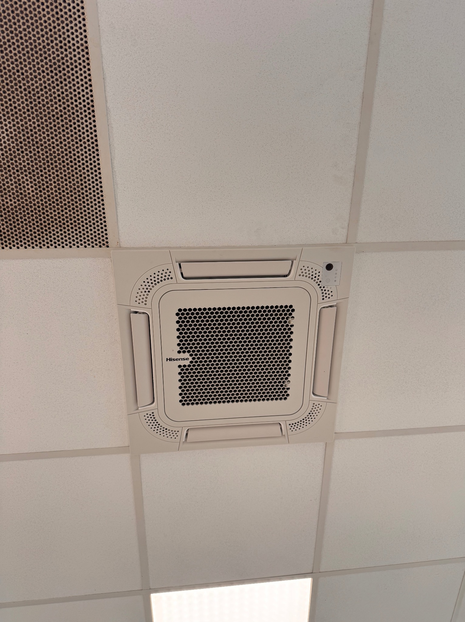 Klimatyzator kasetonowy Hisense wmontowany w sufit podwieszany. Widoczna kratka wentylacyjna z otworami w kształcie plastra miodu i panel sterowania.