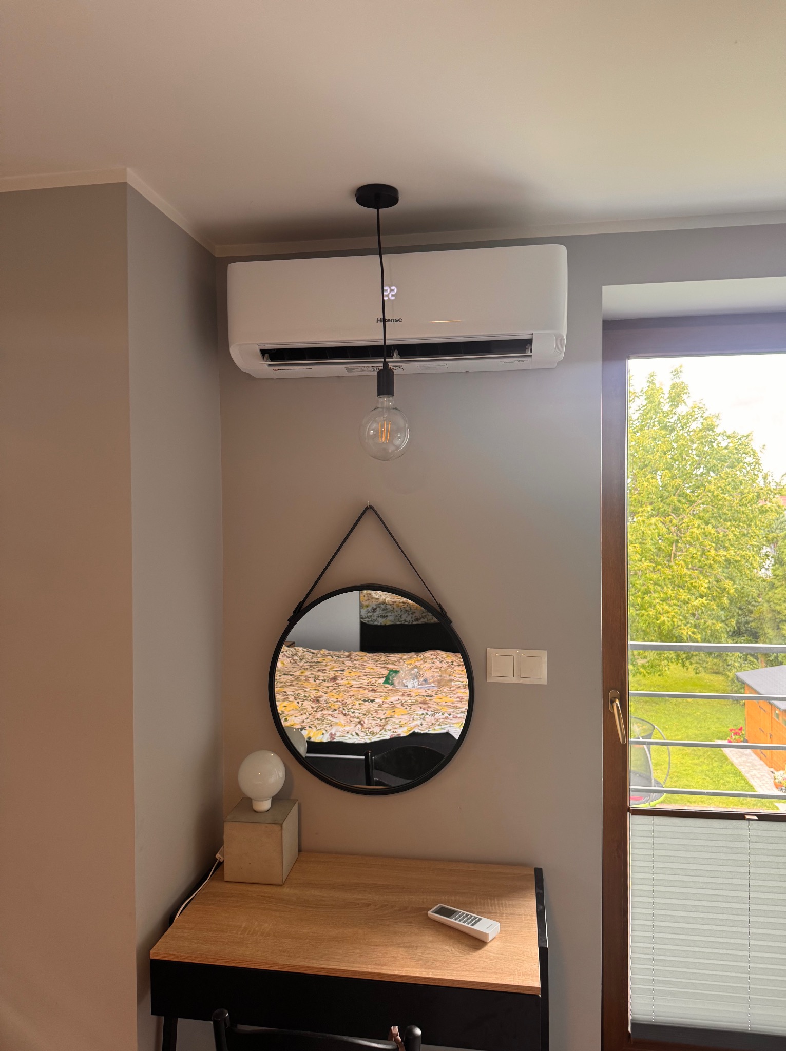 Biały klimatyzator ścienny Hisense w nowoczesnym wnętrzu, zamontowany nad okrągłym lustrem i biurkiem. Widoczny pilot do klimatyzacji.