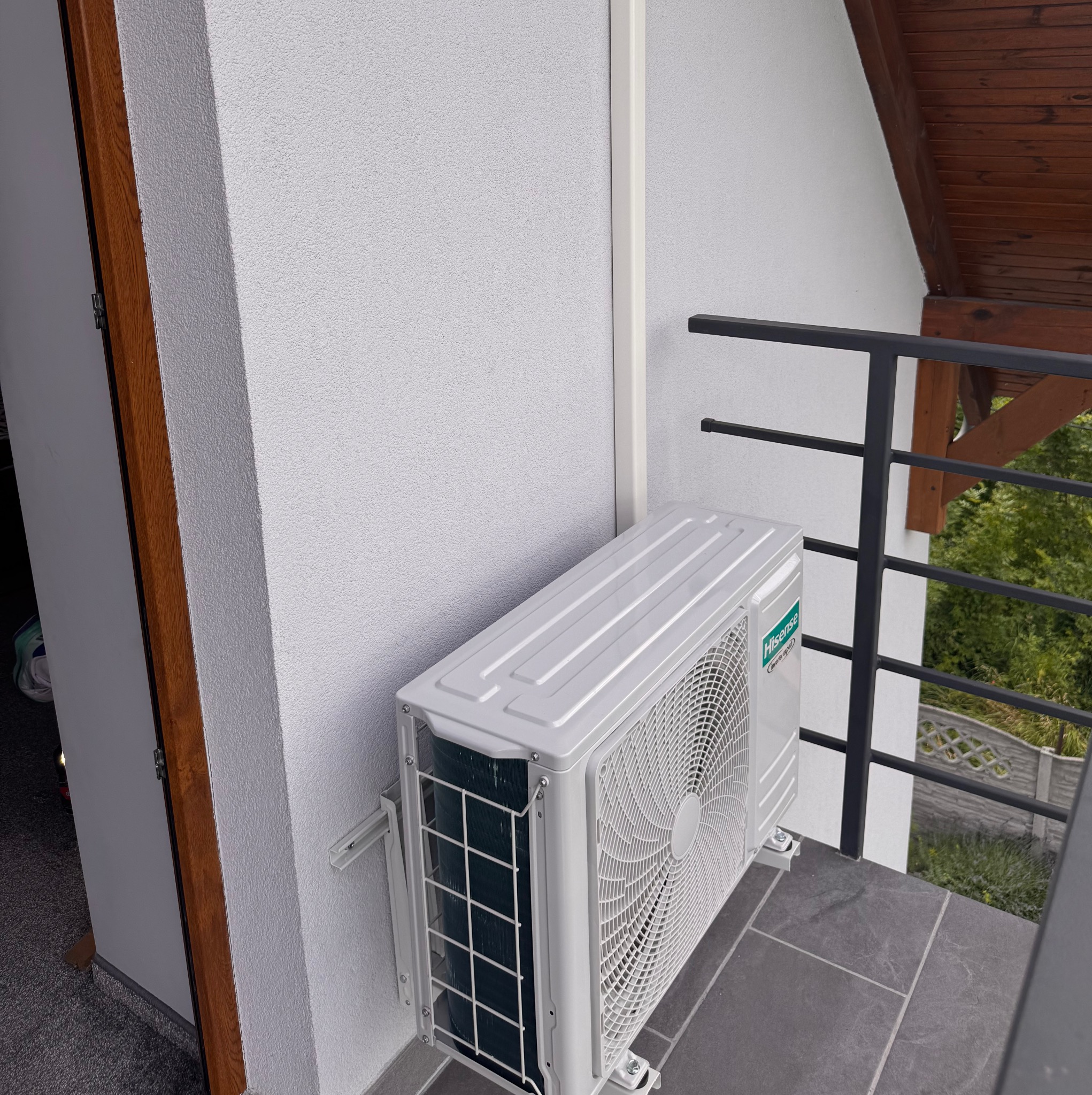 Biała jednostka zewnętrzna klimatyzacji Hisense zamontowana na szarej podłodze balkonu, widok z boku, z fragmentem balustrady i drewnianego zadaszenia.