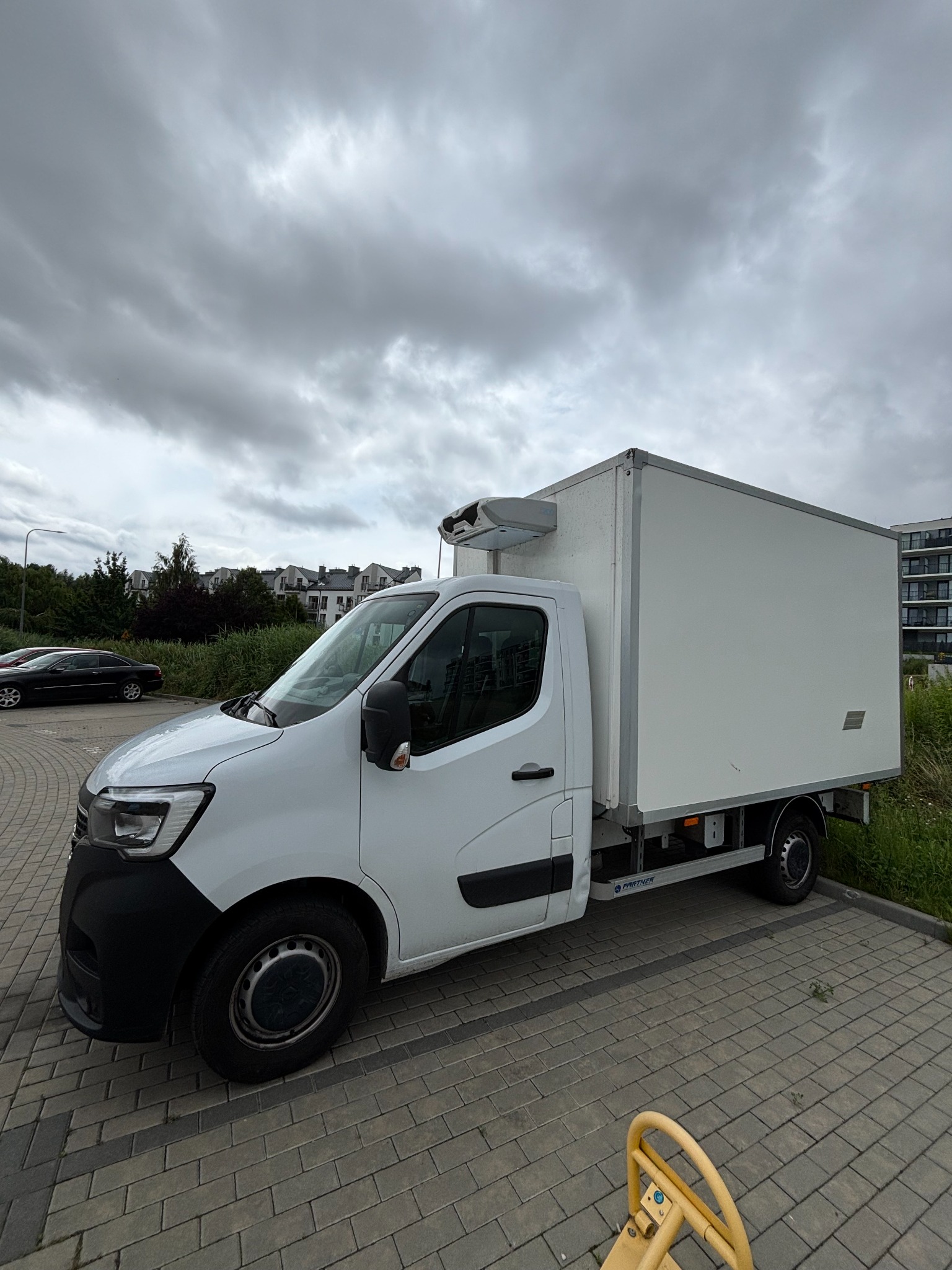 Biały bus chłodnia na parkingu z kostki brukowej, widok z boku, pochmurne niebo w tle.